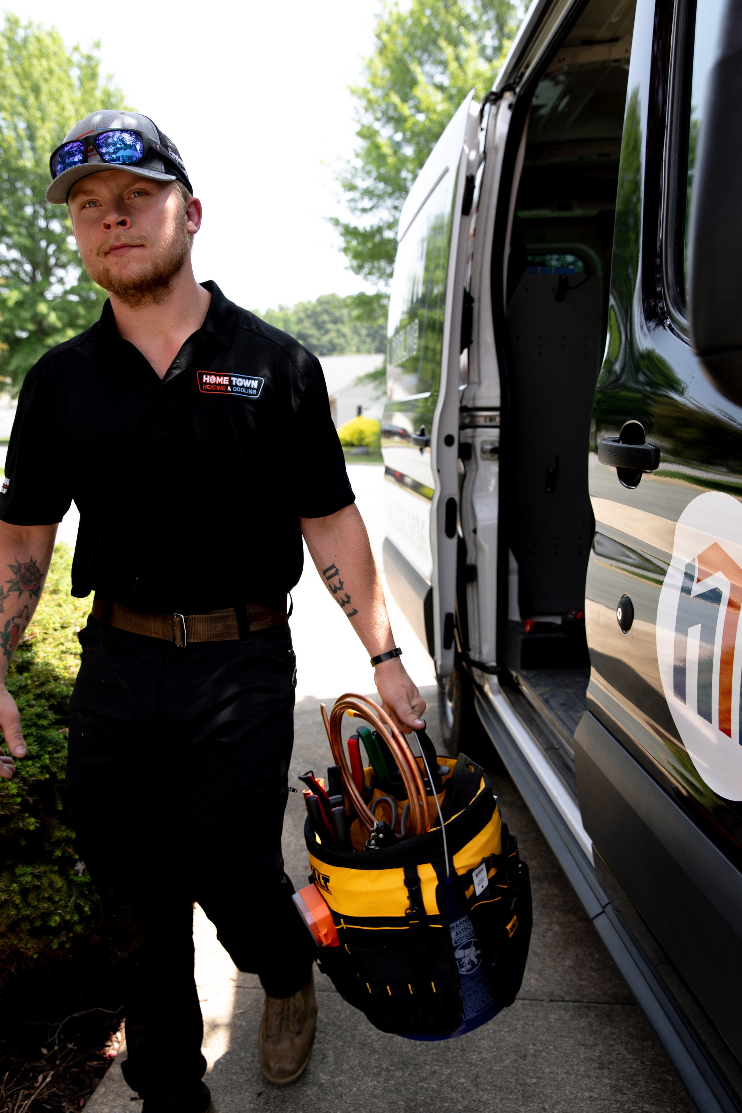 A handyman wearing a black uniform with a logo, sunglasses on his cap, and tattoos on his arms, carrying a bag filled with tools, next to a black van on a sunny day.