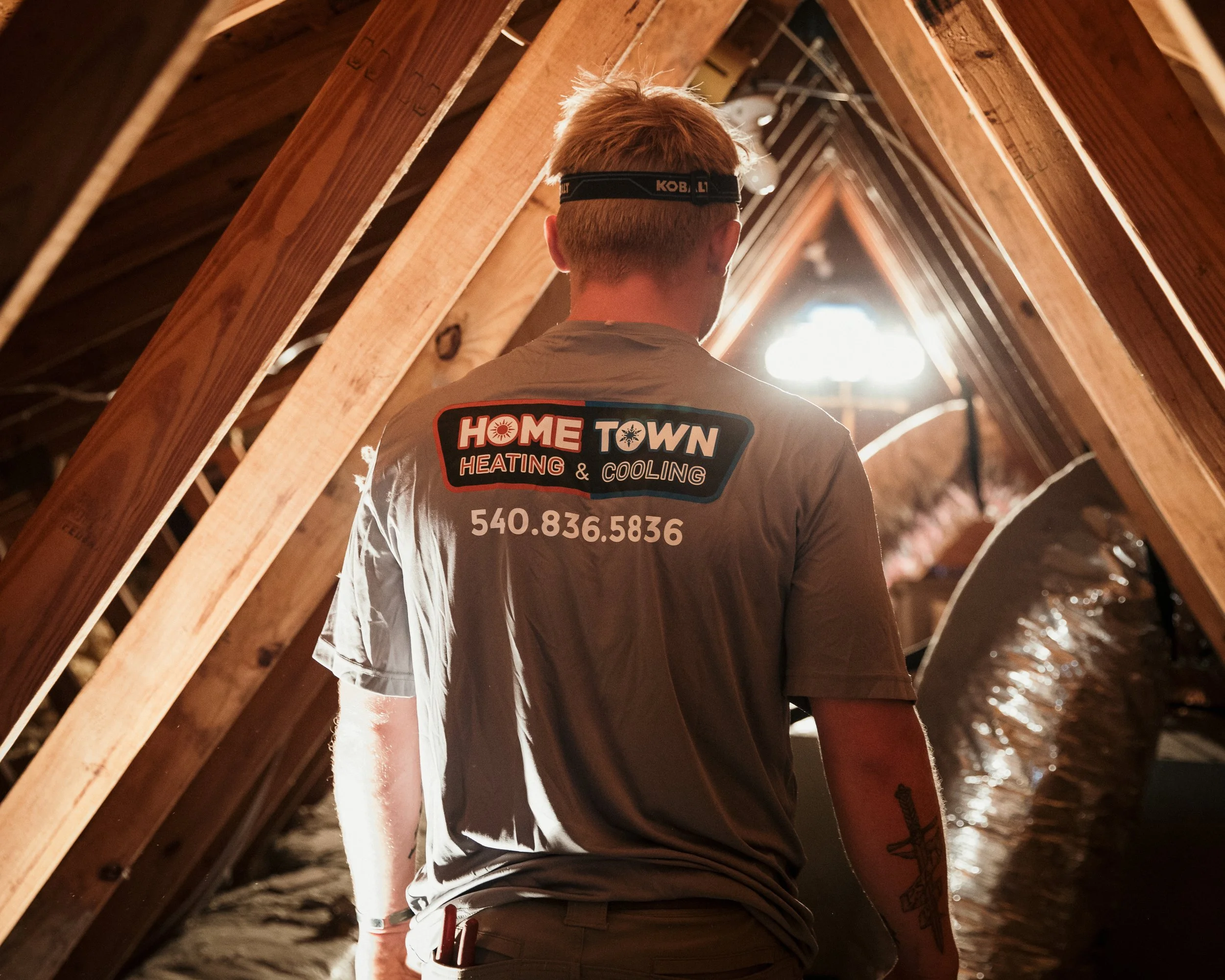 A technician from Home Town Heating & Cooling working in an attic with wooden beams and insulation, facing away from the camera.