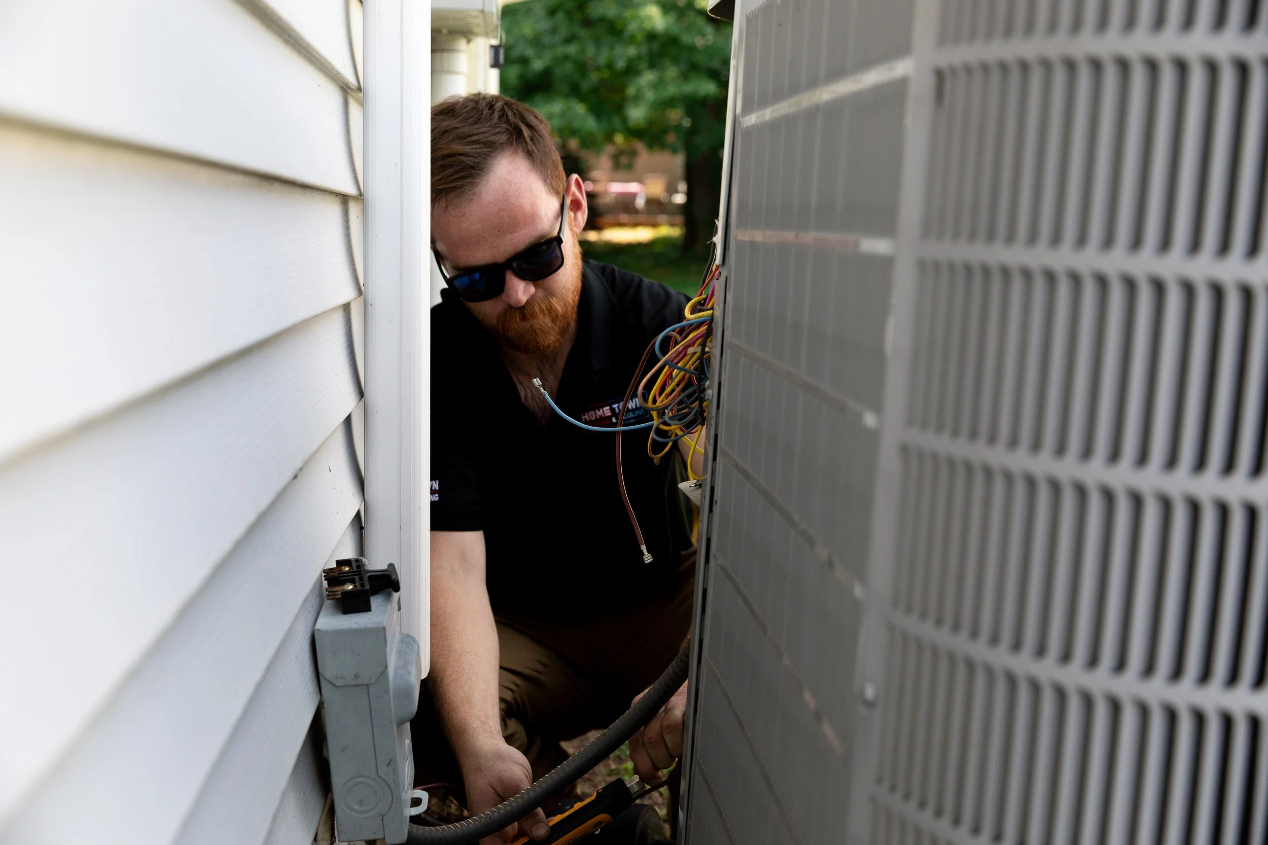 A man with a beard and sunglasses working on an air conditioning unit outside a house.