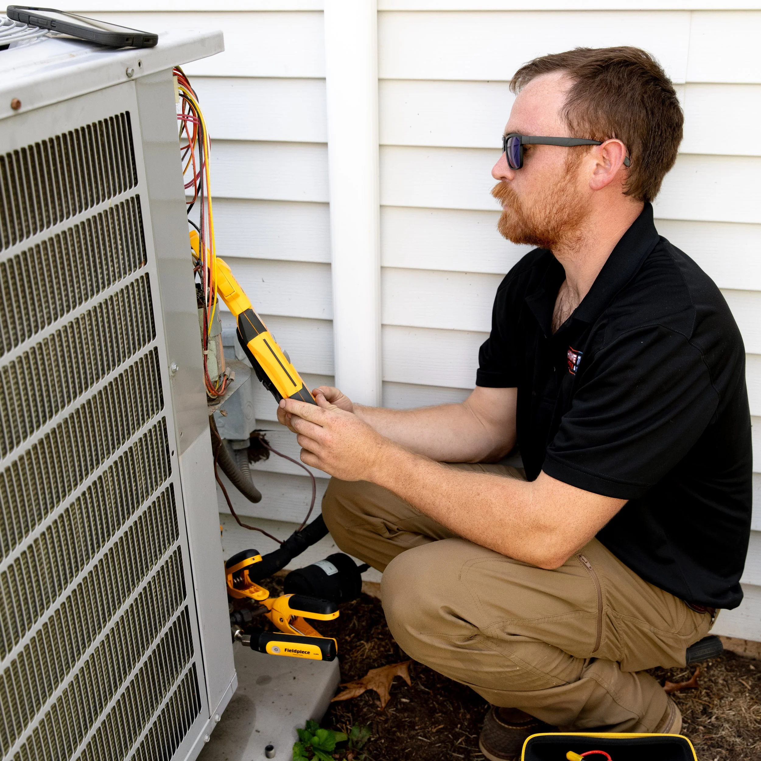 An HVAC Technician working on an outdoor air conditioning unit, using a multimeter, with tools nearby, in front of white siding.
