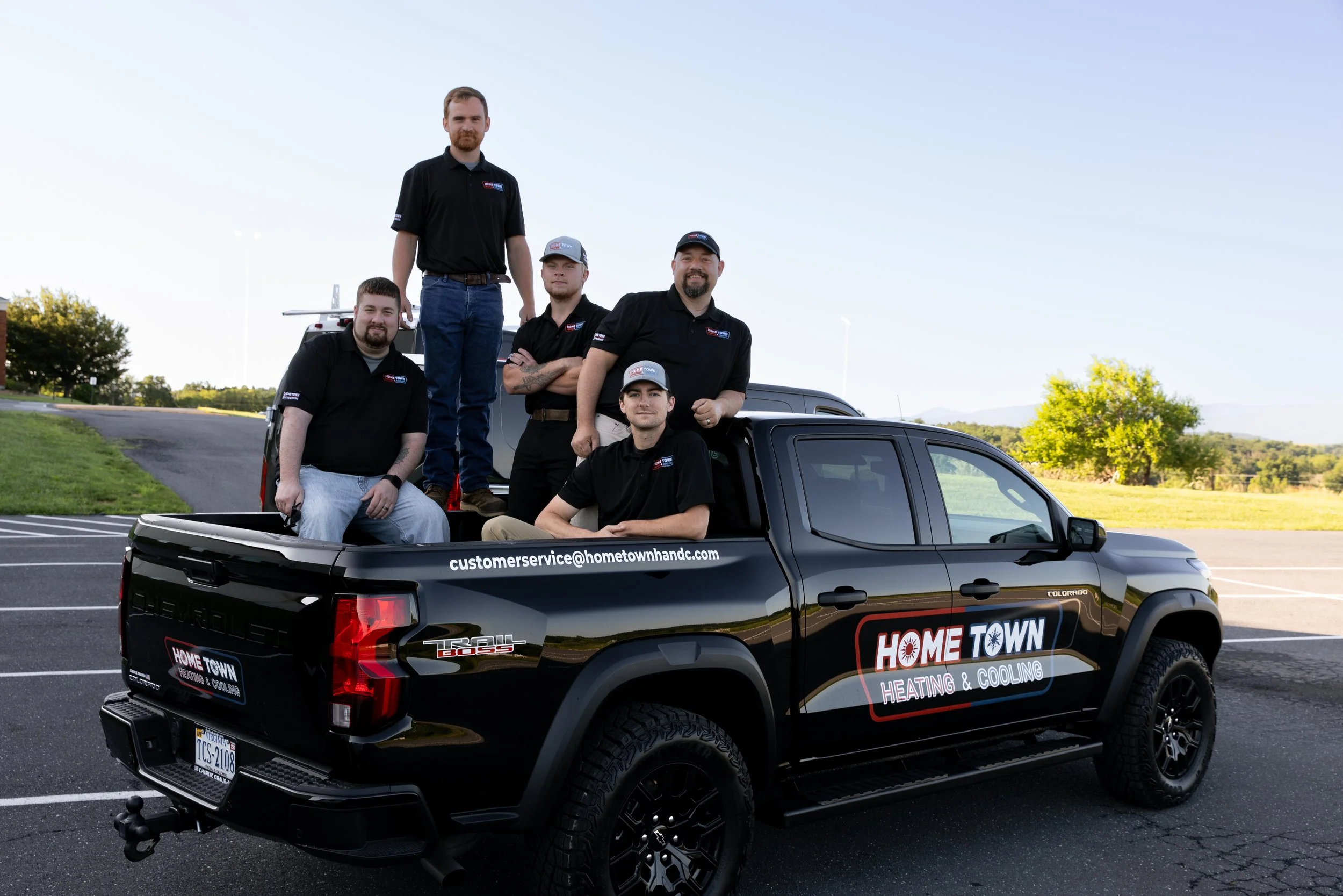 Group of five men in black Home Town Heating & Cooling uniforms posing around a black pickup truck in a parking lot with a grassy field and trees in the background.