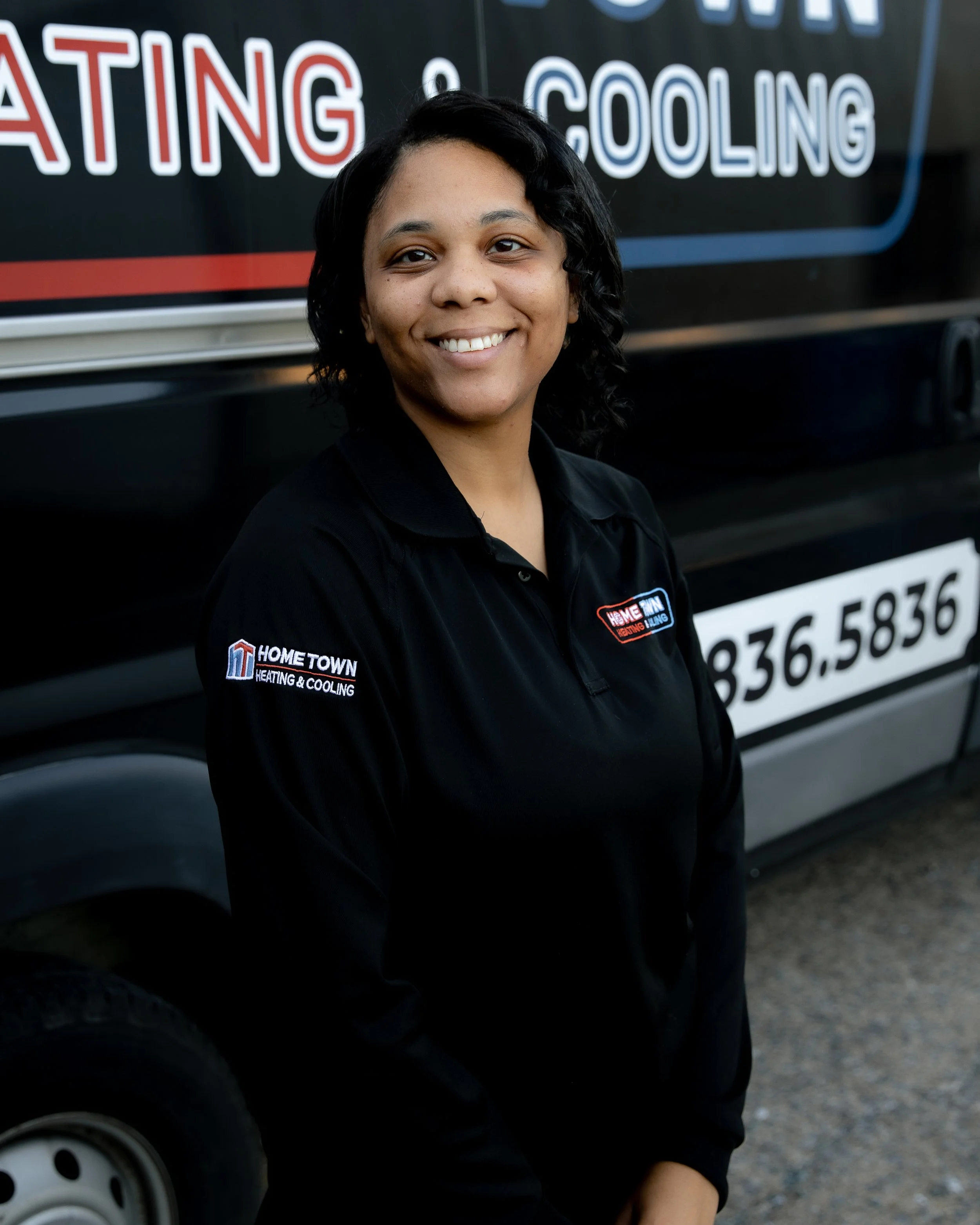 A smiling woman wearing a black shirt with the logo 'Hometown Heating & Cooling' standing in front of a black service van with the same logo and a phone number.