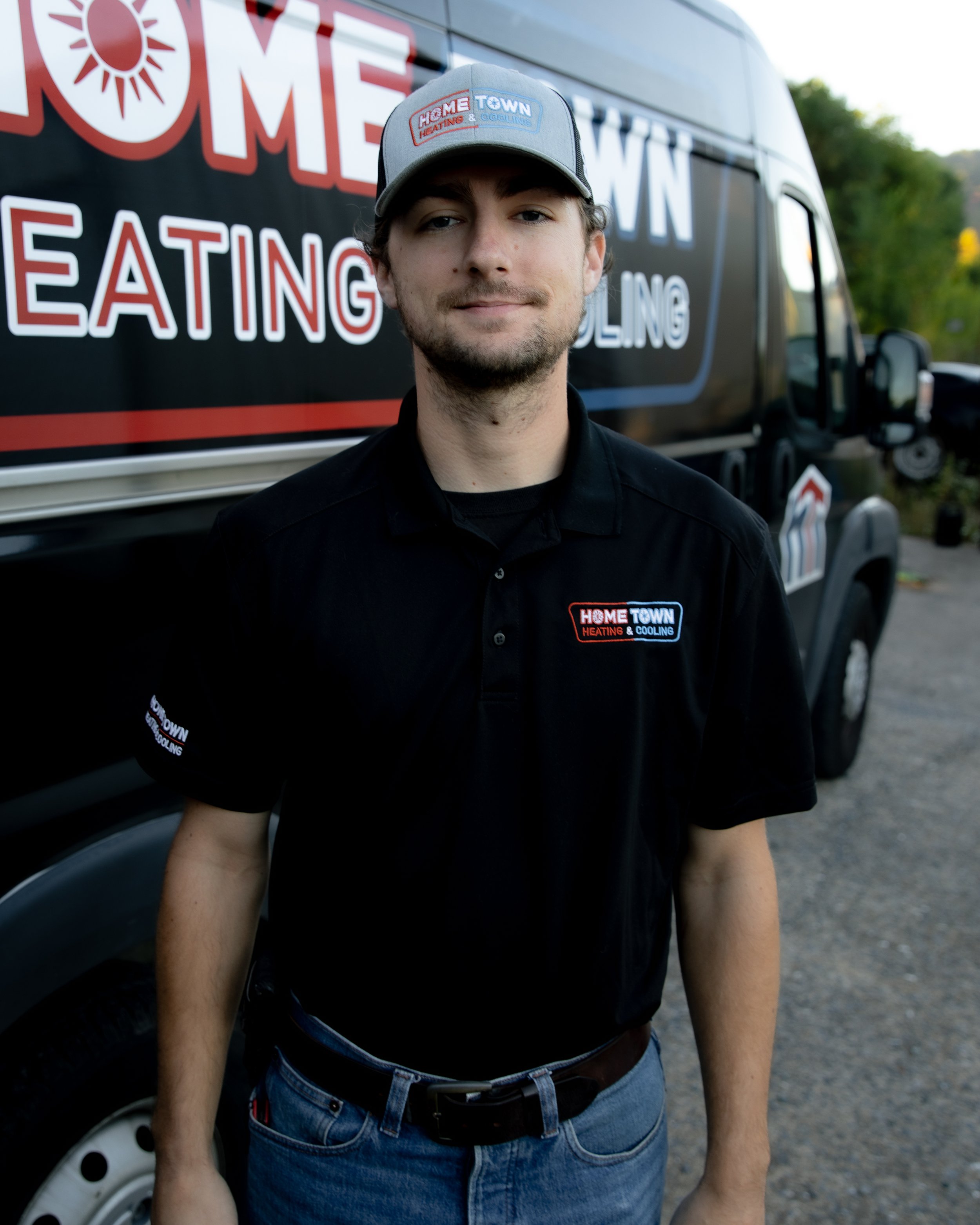 A young man in a black uniform with a 'Home Town Heating & Cooling' patch stands outside in front of a truck with similar branding. He wears a gray cap with the same company logo.