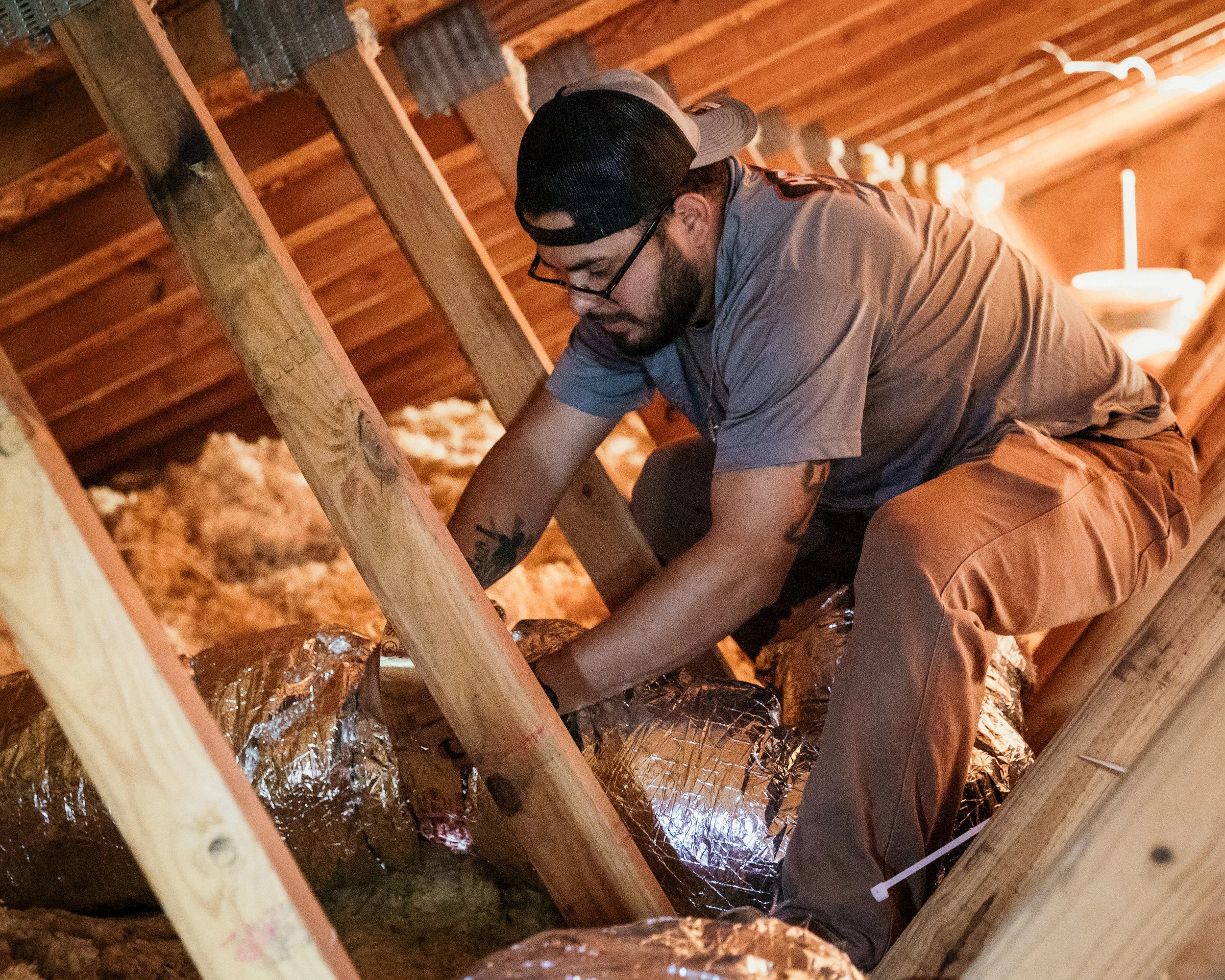 A man working inside an attic, installing or inspecting insulation wrapped in aluminum foil among wooden beams.