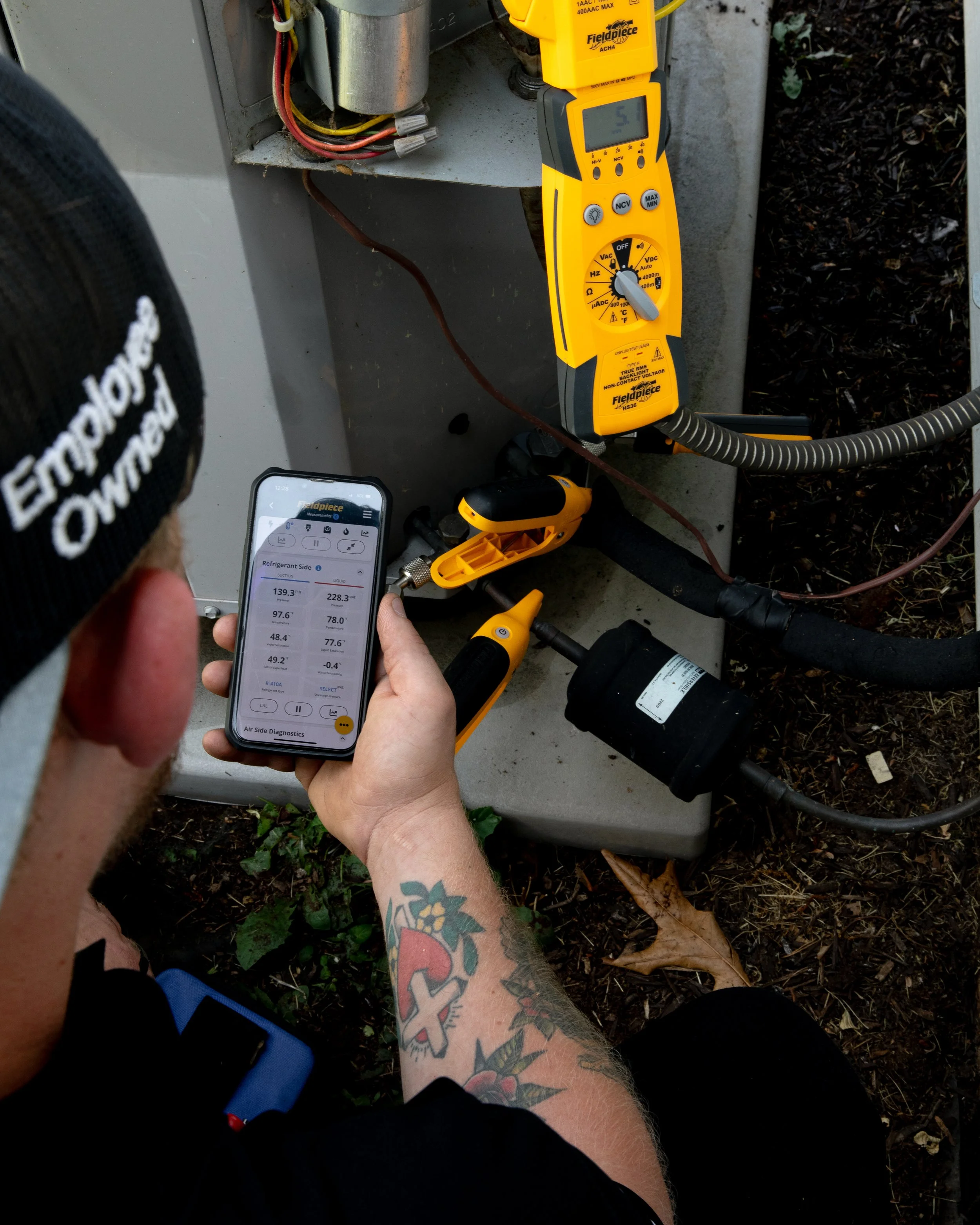 Technician using a digital multimeter and an infrared thermometer to diagnose an HVAC unit outdoors.