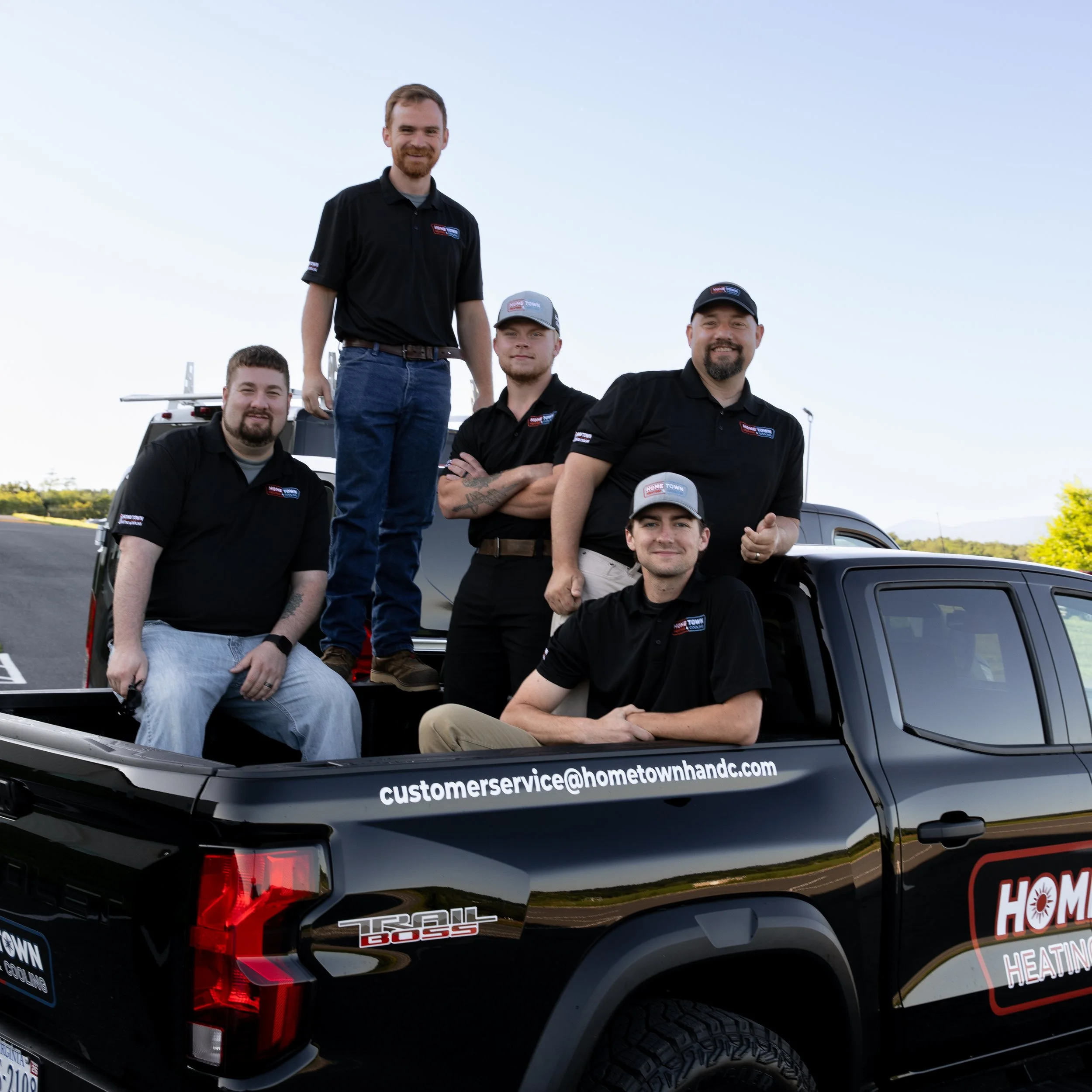 A team of five men posing on and around a black pickup truck with the company logo 'Hometown Heating & Cooling' on the side. The truck has a contact email visible. They are outdoors under a clear sky, with some trees in the background.