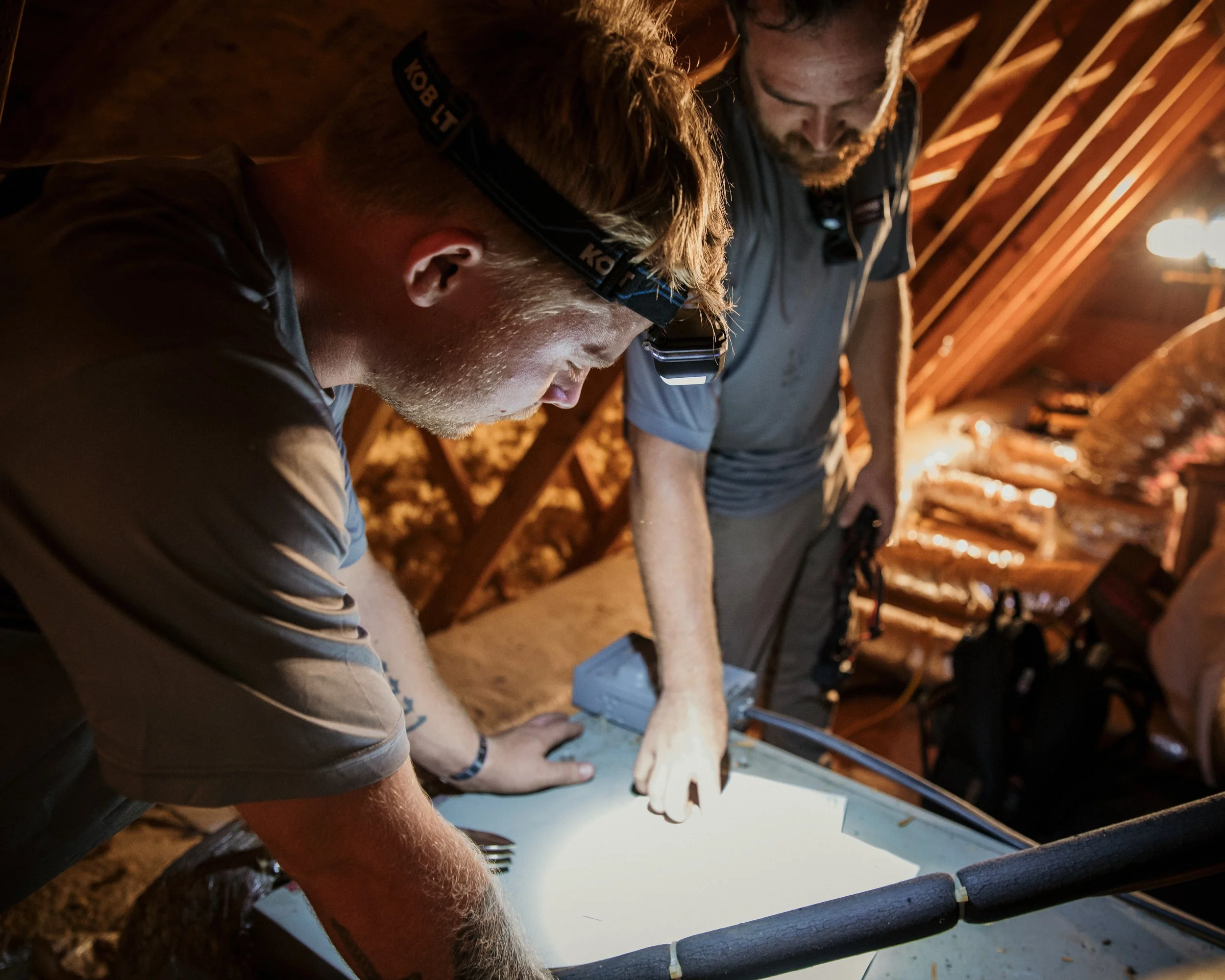 Two men working together inside a wooden attic, examining or working on a flat surface with a bright light illuminating their workspace.