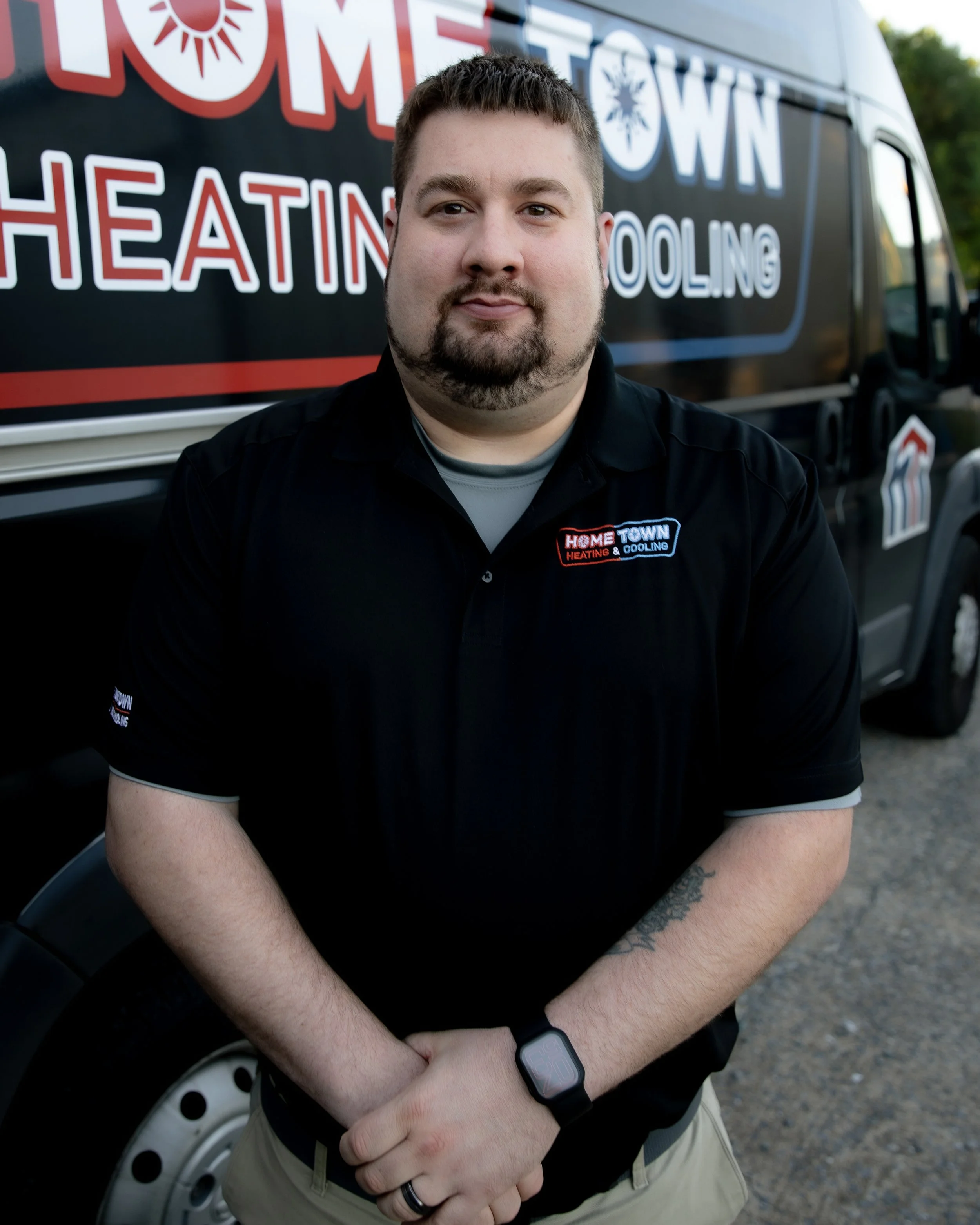 Man wearing a black shirt with a company logo, standing in front of a service van with company branding for "Home Town Heating & Cooling".