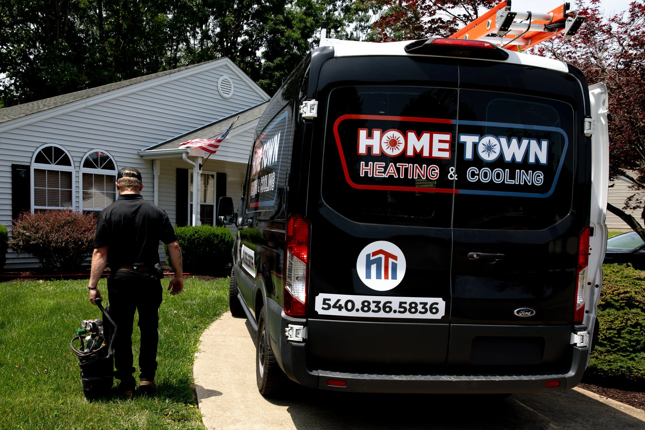 A technician walking away from a service van parked in front of a house, carrying a toolbox in a residential neighborhood. The van has branding for Home Town Heating & Cooling, including a phone number and logo.