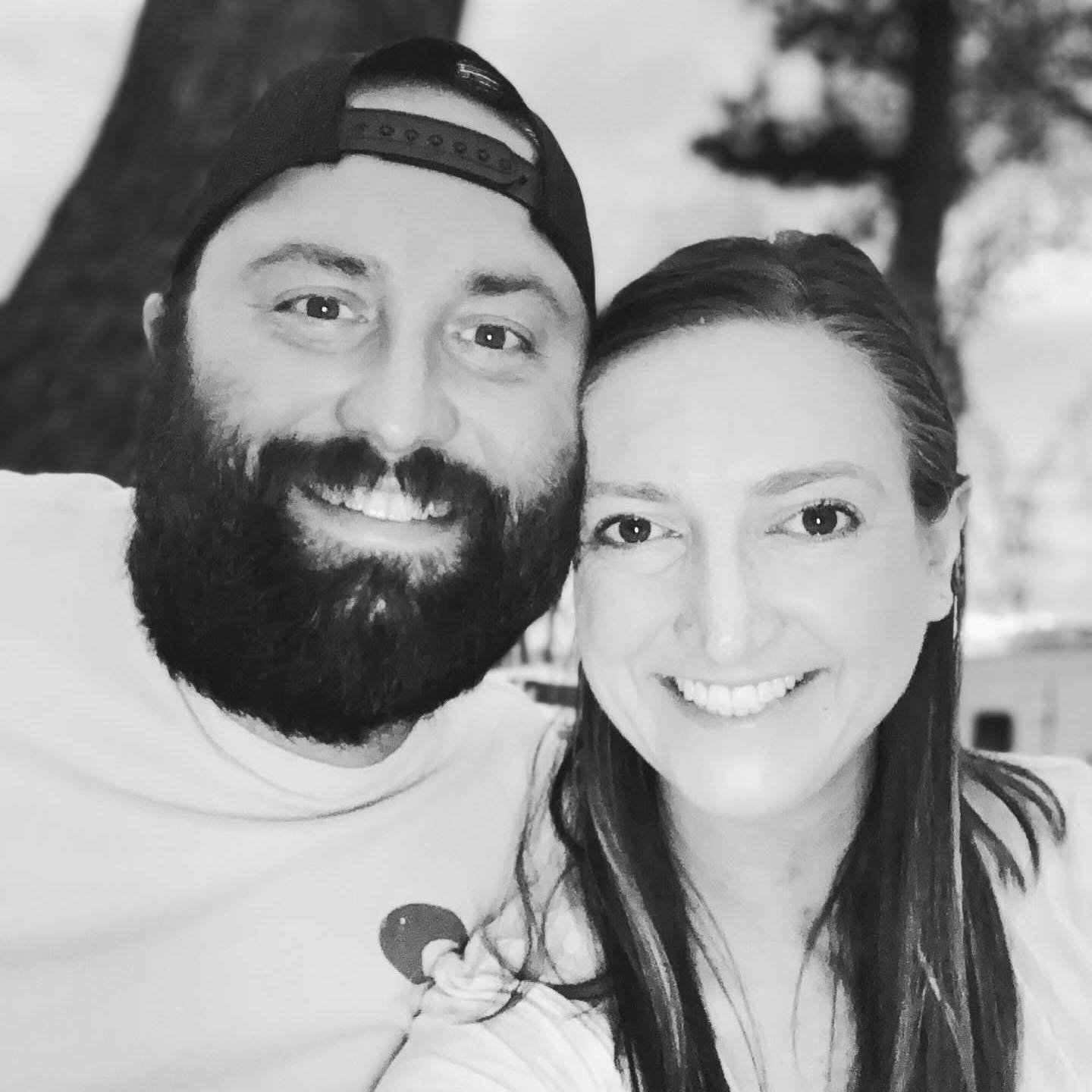 A smiling man with a beard wearing a baseball cap backwards and a woman with long hair, both smiling in a close-up selfie outdoors.