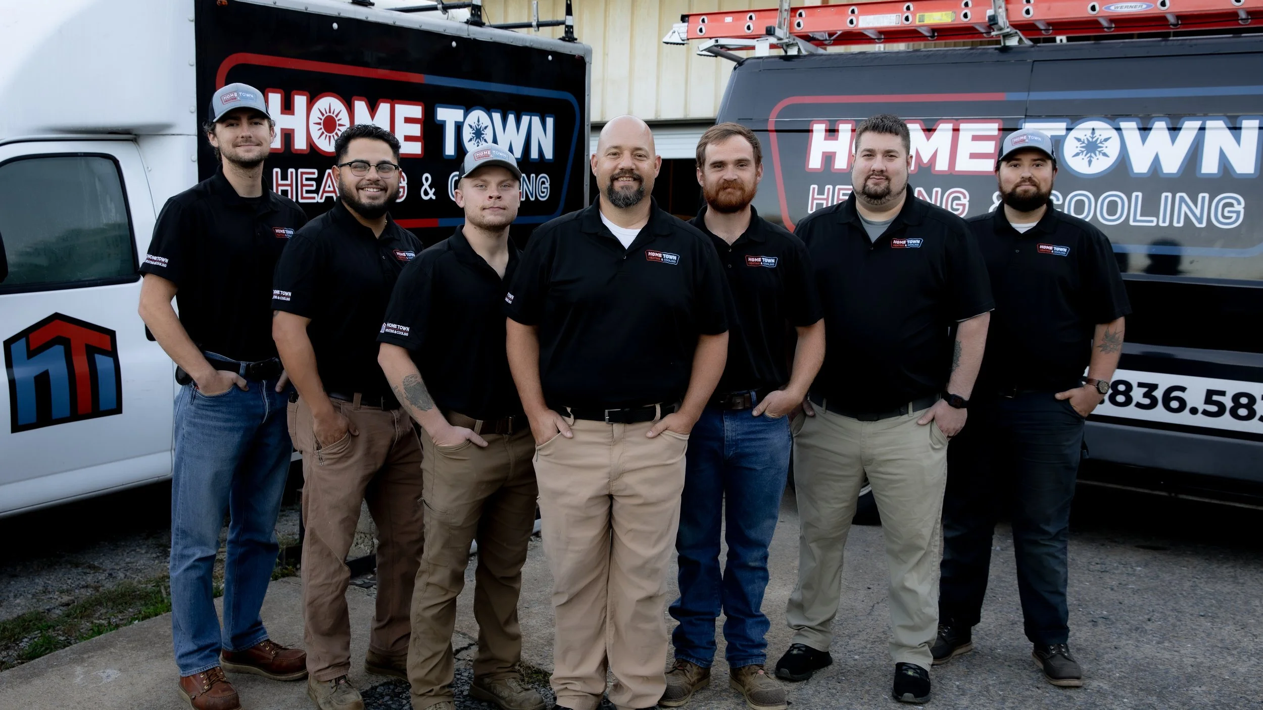 Group of seven men standing side by side in front of service trucks with 'Home Town Heating & Cooling' signage, all wearing black polo shirts with company logo. Some have tattoos and glasses, and wear caps and casual pants.