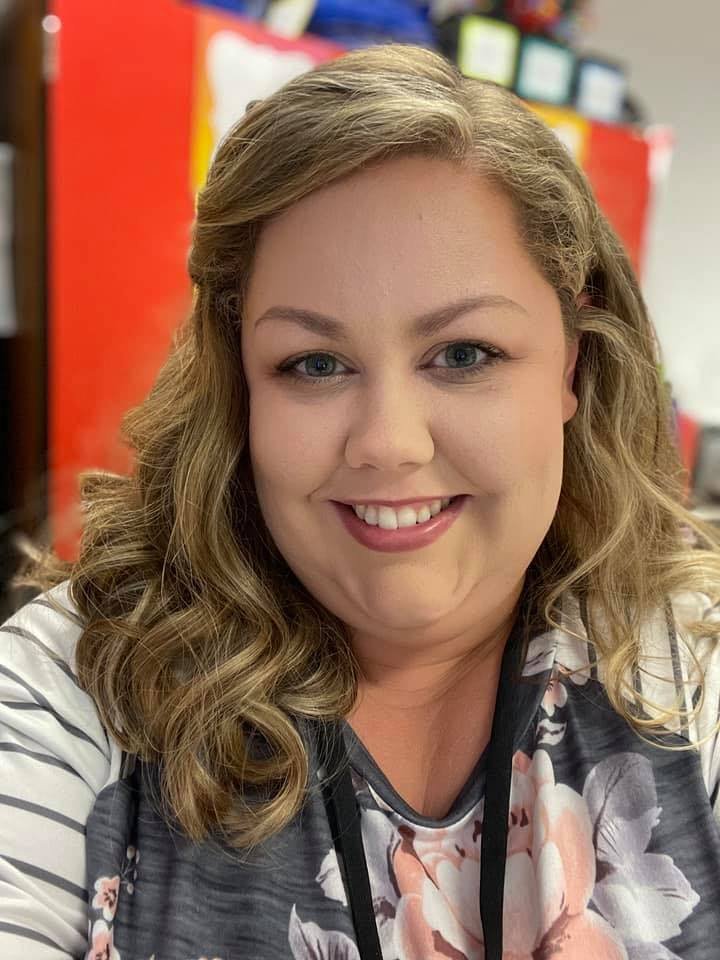 A woman with wavy blonde hair smiling at the camera in a classroom or office setting.