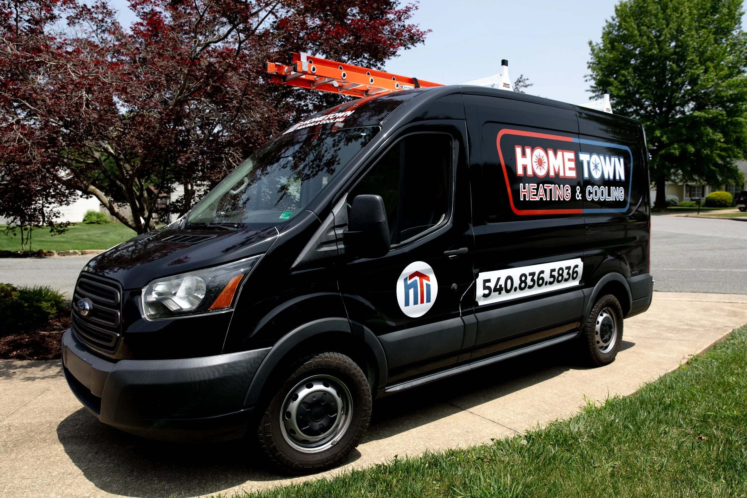 Black service van parked on a driveway with company branding, trees in the background, and a ladder on top.
