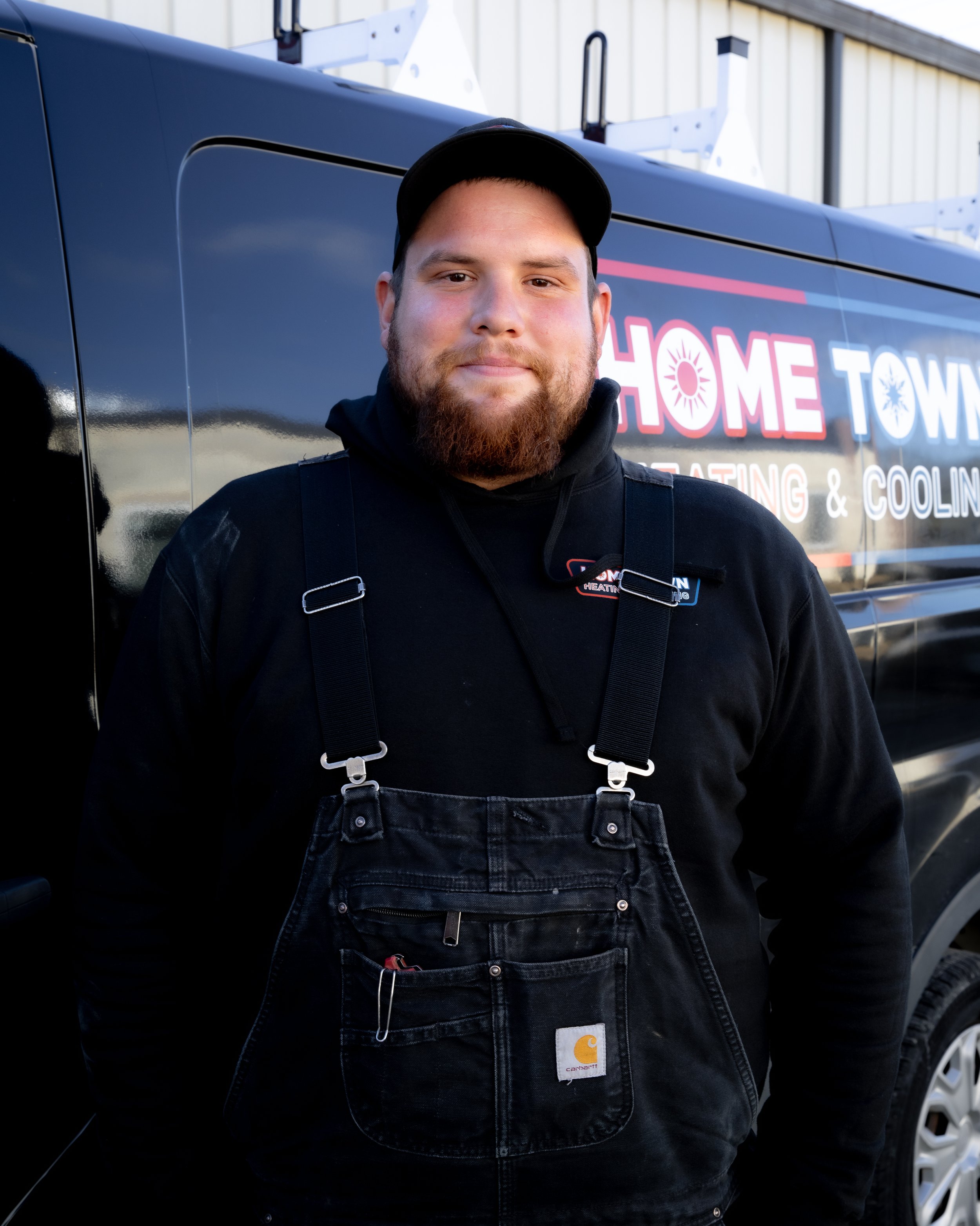A man with a beard and mustache wearing a black Carhartt bib and a black hoodie, standing in front of a black service van with the logo and signage for a heating and cooling company.