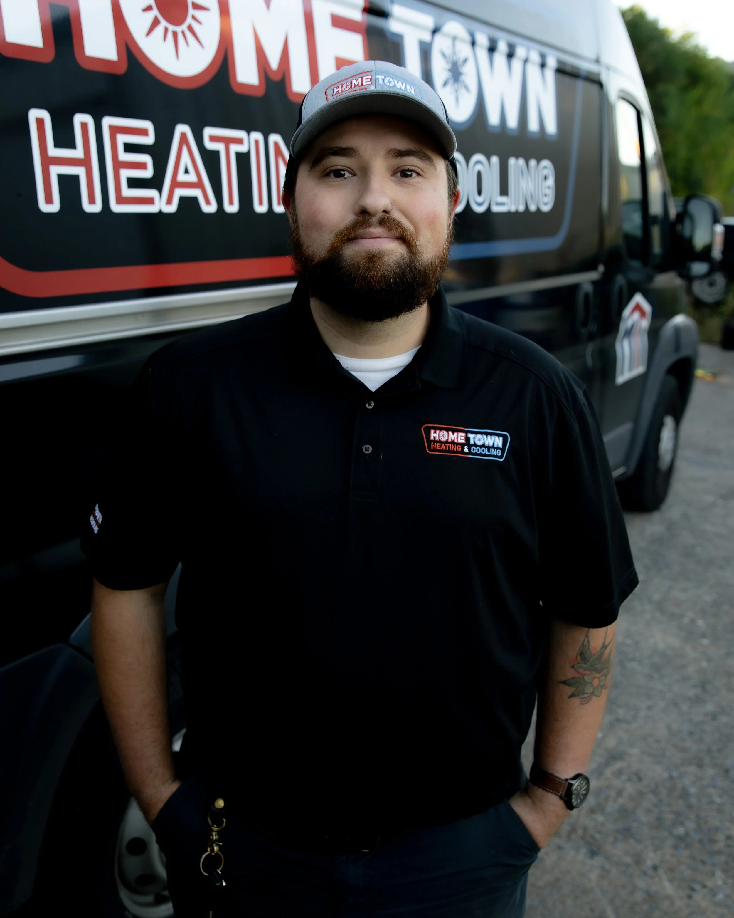 A man stands outdoors in front of a black vehicle with a logo for HomeTown Heating & Cooling. He is wearing a black polo shirt with the company logo, a gray cap with the company name, and has a tattoo on his right forearm.