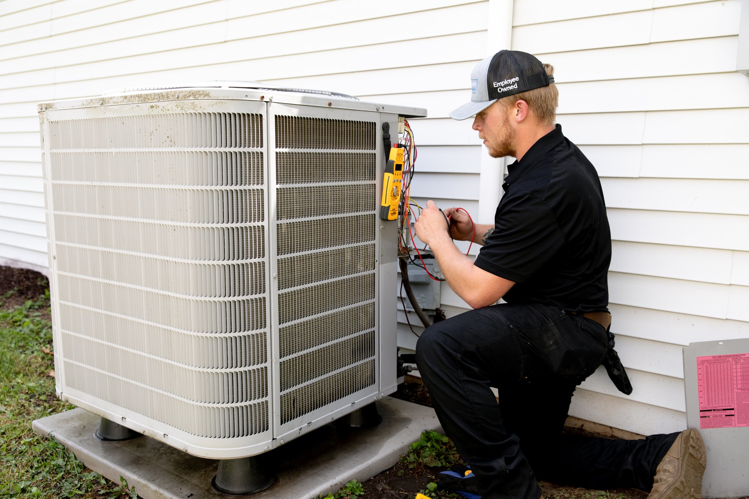 A technician kneeling outside, working on a central air conditioning unit with a multimeter and wires, in front of a white house siding.