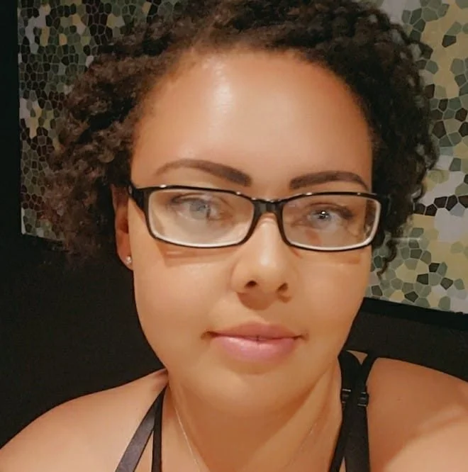 Close-up photo of a woman with short, curly brown hair, wearing black-framed glasses, light makeup, and a black strap top, sitting in front of a patterned wall.