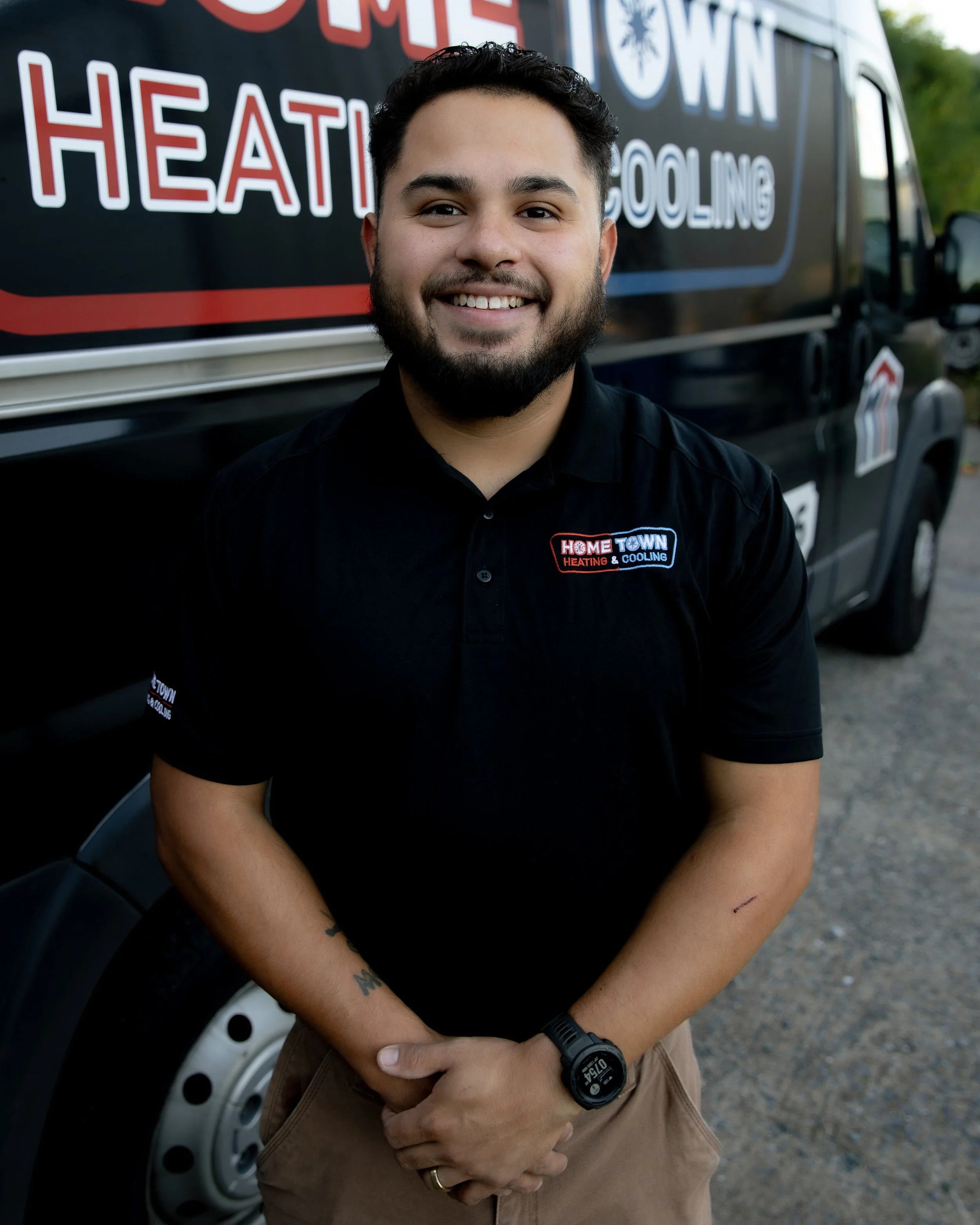 A smiling man wearing a black polo shirt with a company logo, standing in front of a company vehicle with similar branding.