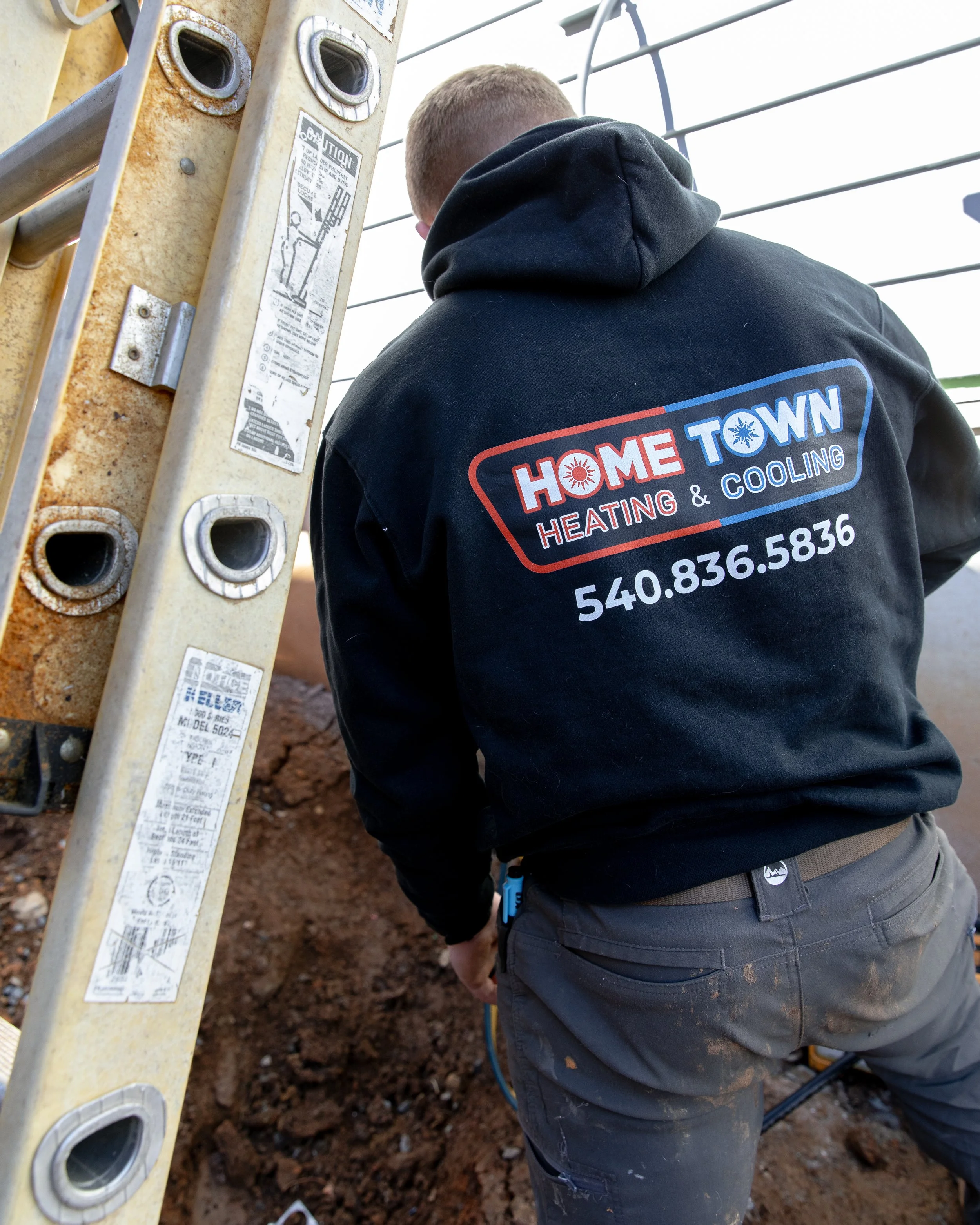 A worker from Home Town Heating and Cooling is working outdoors near a metal ladder and a wire fence. The worker is wearing a black hoodie with the company's logo and phone number on the back, and is bent over working on the ground.