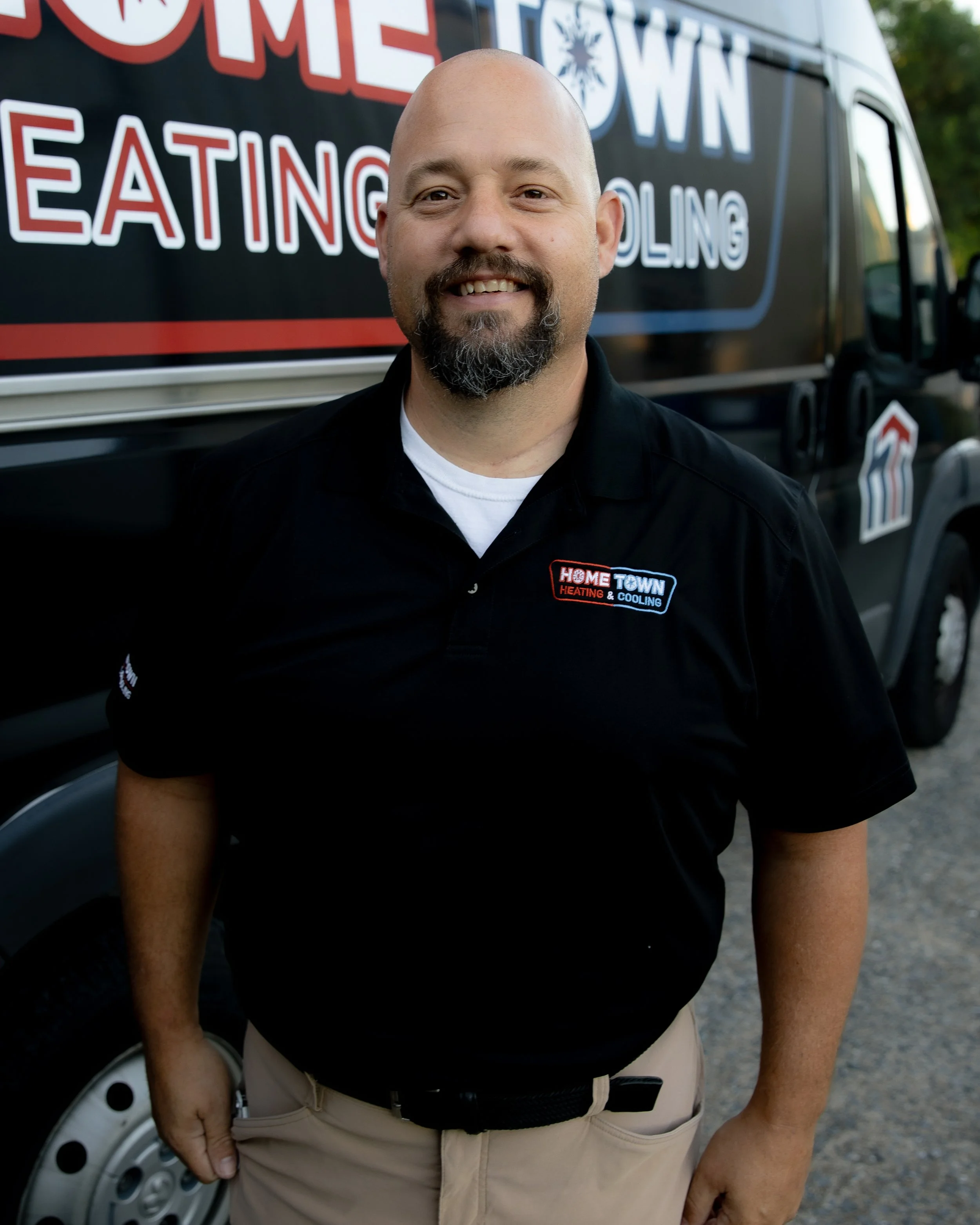 Man standing in front of a service van with the company logo, wearing a black polo shirt with the same logo, smiling at the camera.