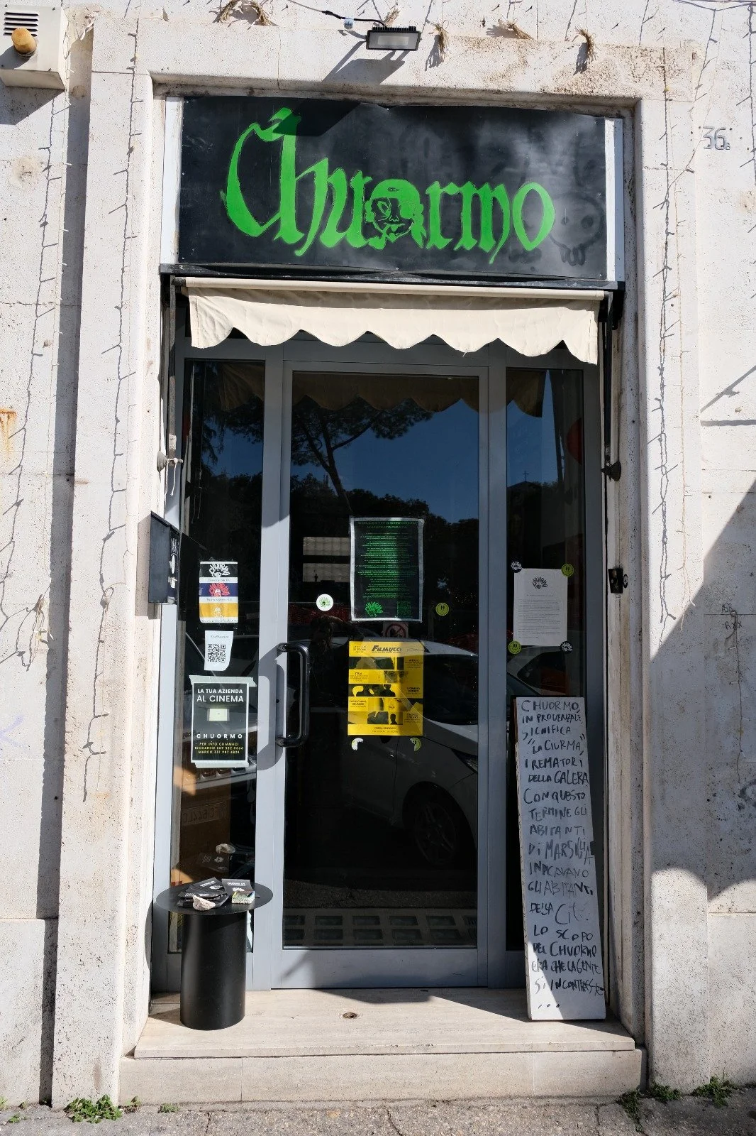 Front entrance of a store with a black signboard with green gothic text reading 'Churmo', a glass door, a white canopy, and handwritten signs on a white board outside.