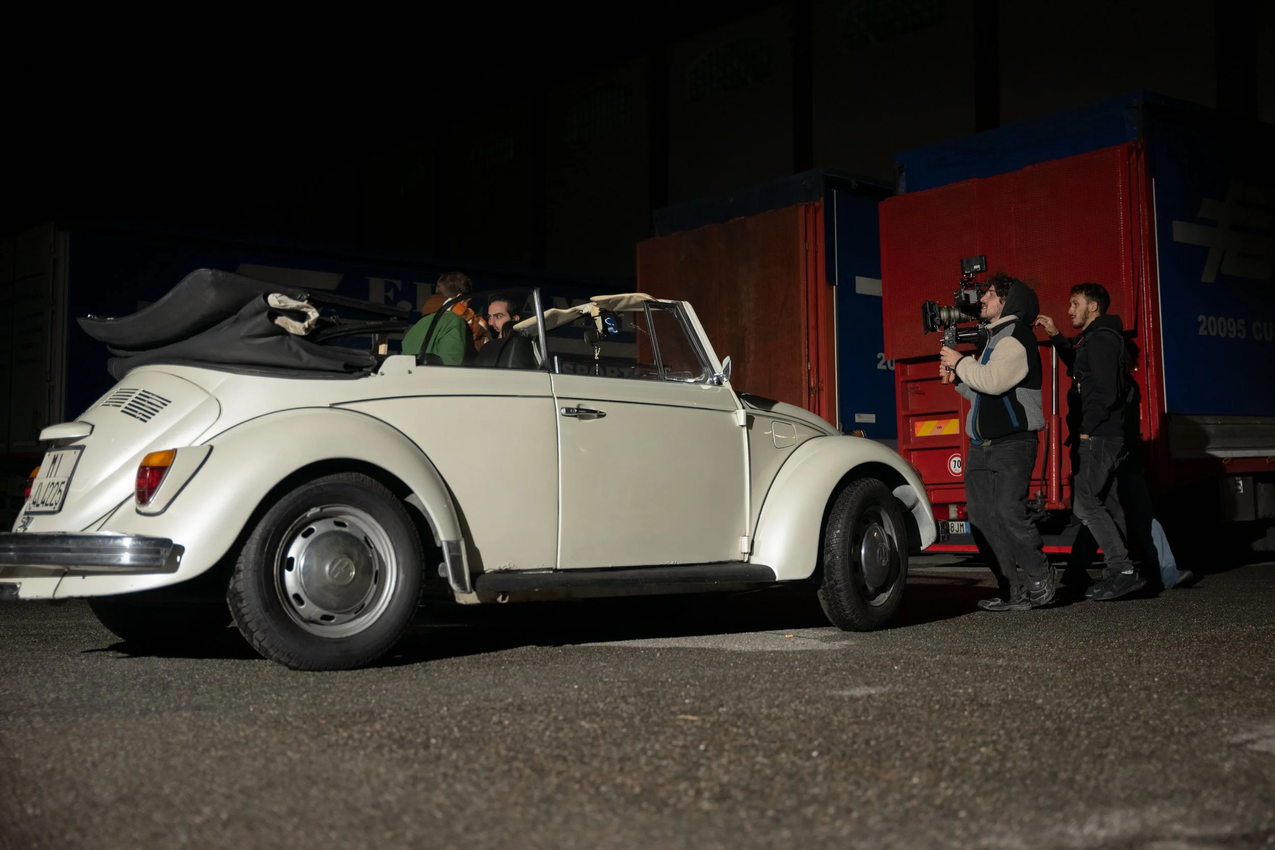 Night scene of a vintage white convertible car with a black soft top, parked in front of large red and blue semi-truck trailers. Two people sit inside the car, while three crew members are filming it; one is holding a camera, and another is behind him.