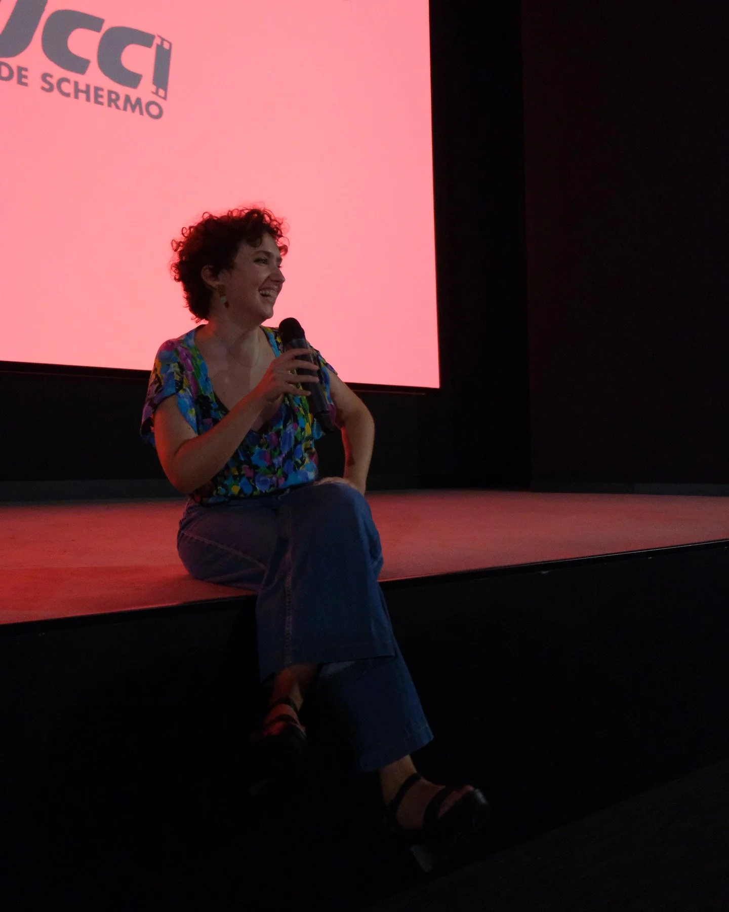 A woman sitting on a stage with a microphone, smiling, in front of a pink screen with 'YCCI DE SCHERMO' written on it.