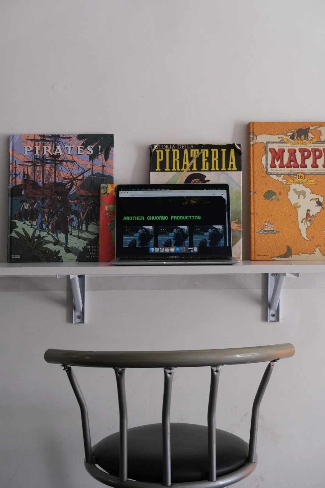 A white shelf mounted on a white wall with four books about pirates and maps, and a laptop displaying green text and a video, positioned above a modern silver and black chair.