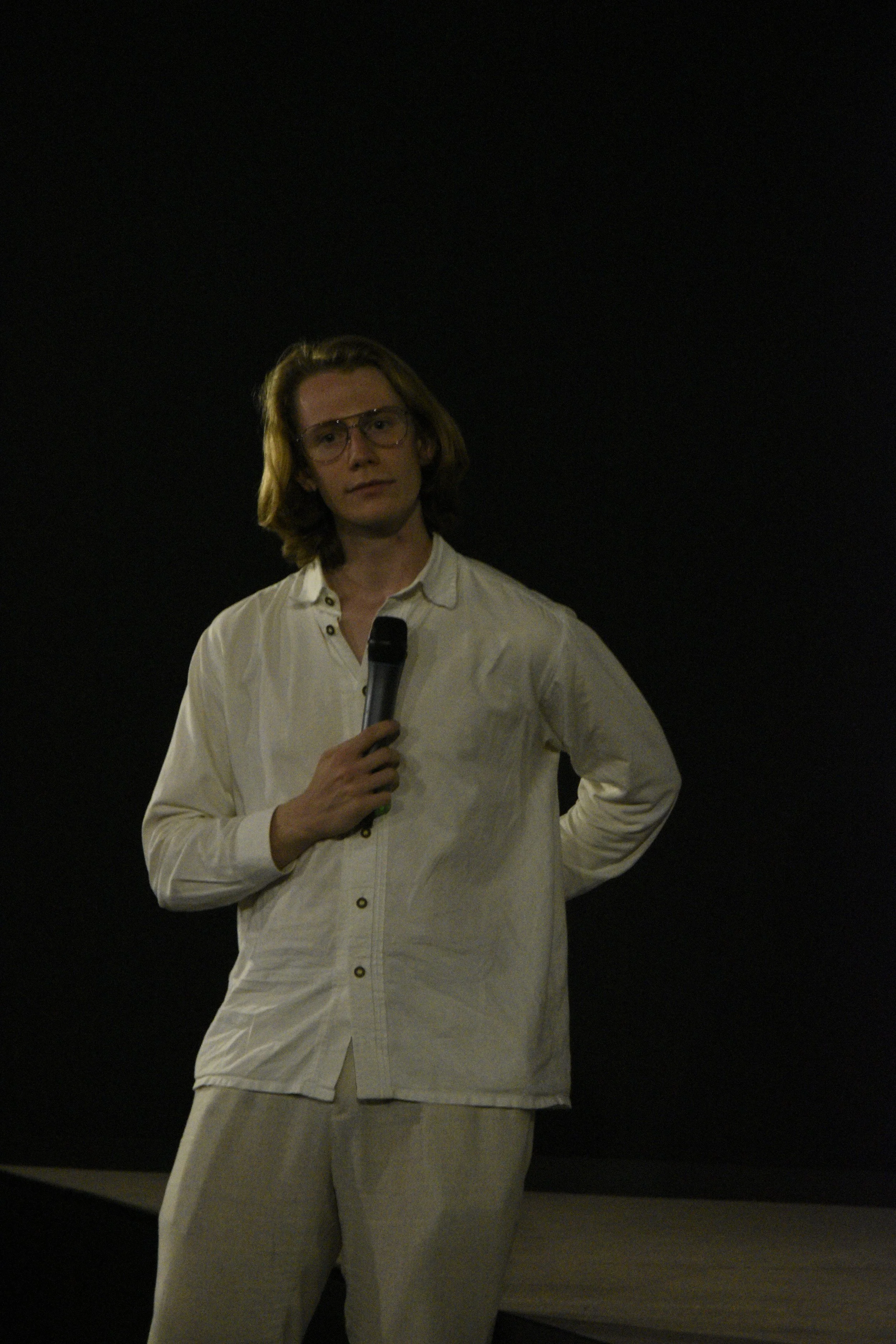 A young man with shoulder-length brown hair, wearing glasses and a white button-up shirt, stands holding a microphone in front of a dark background during a presentation or speech.