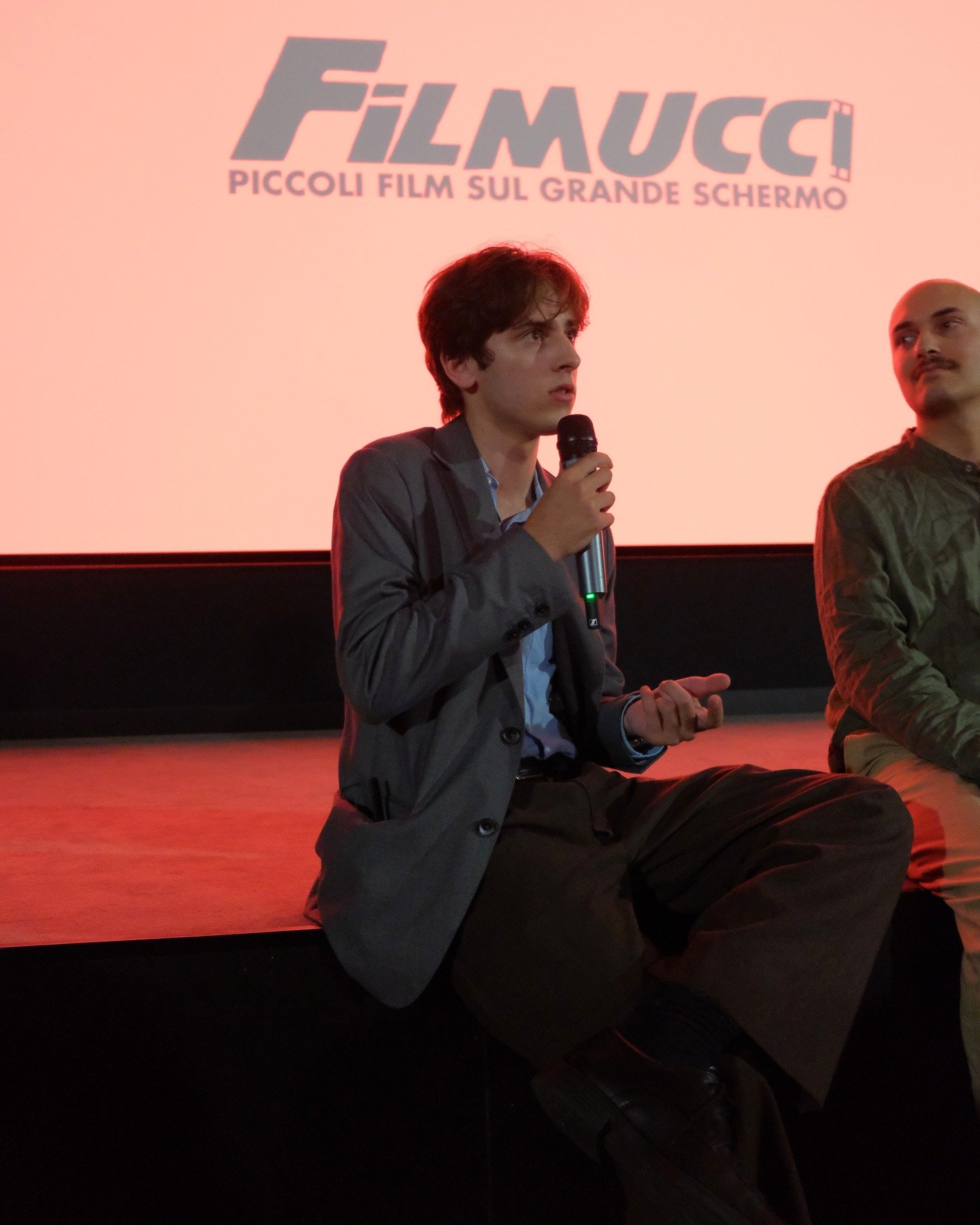A man sitting on a stage with a microphone, in front of a screen displaying the logo 'FILMUCCI' and the tagline 'Piccoli film sul grande schermo'.