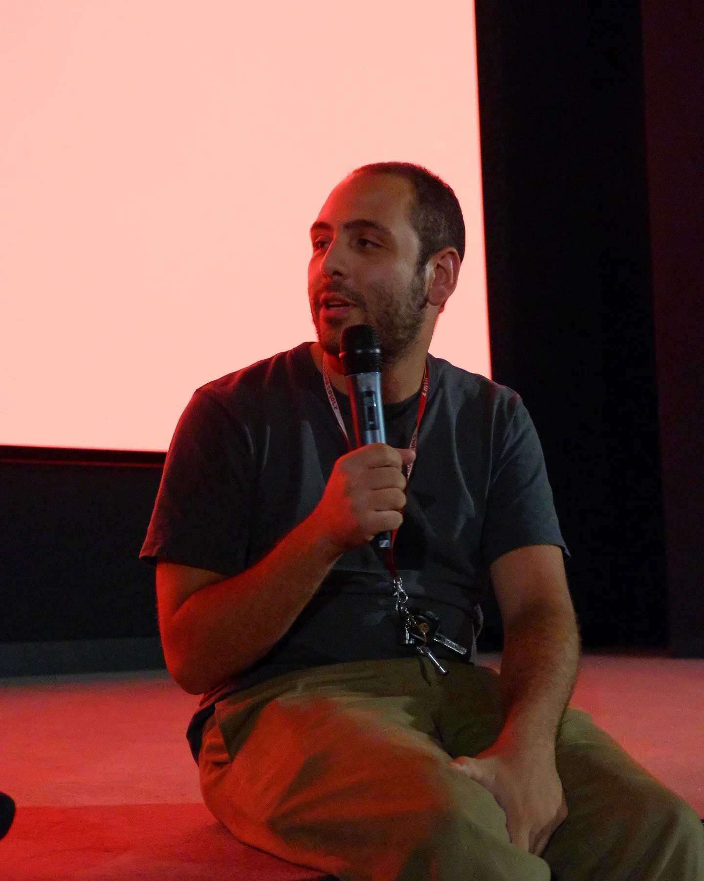 A man sitting on the floor holding a microphone, speaking or answering questions, with a red and black background.