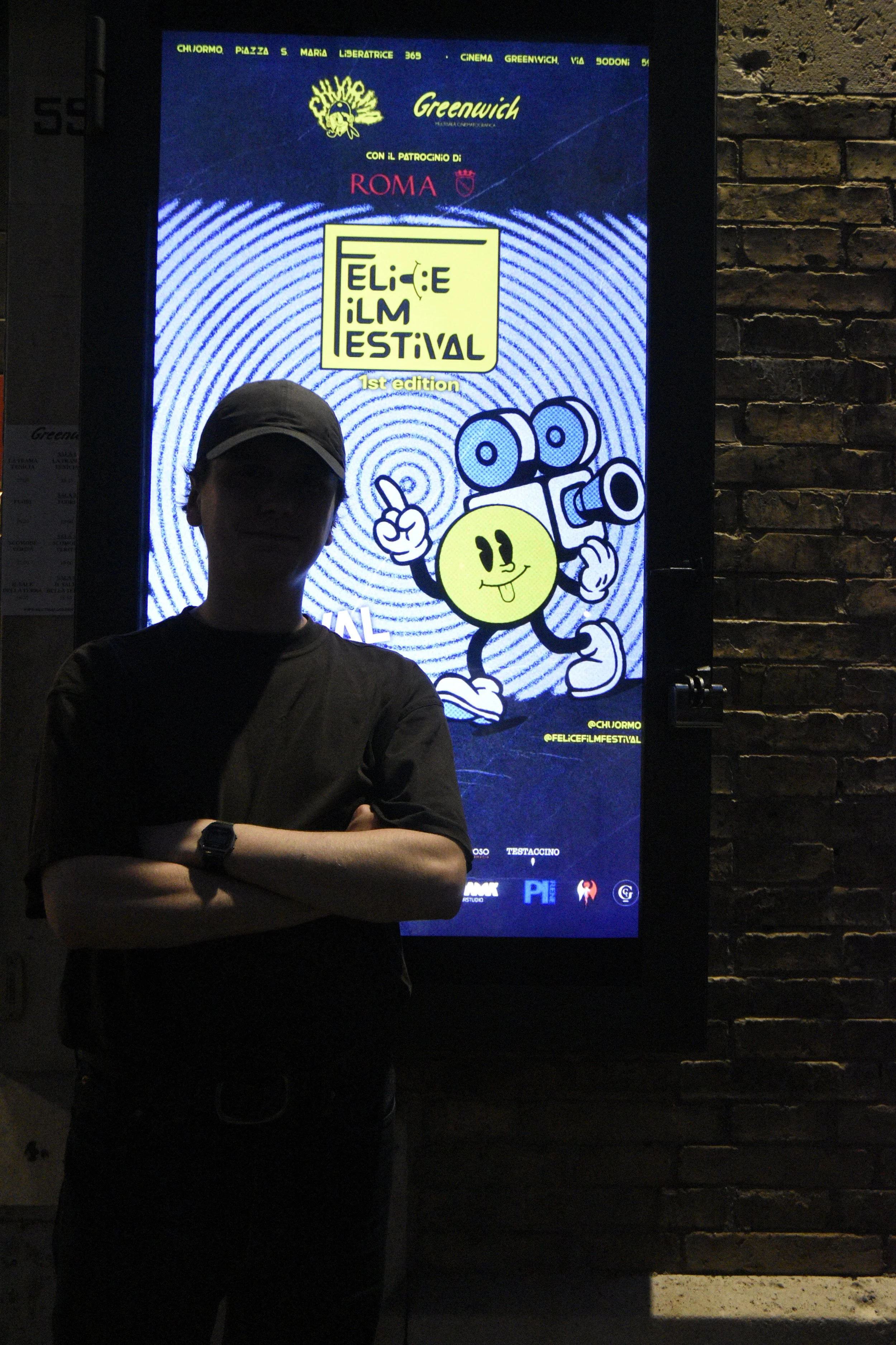 Person wearing a dark shirt and cap standing in front of a lit-up sign for the Felice Film Festival, with a cartoon character holding a camera on the sign.