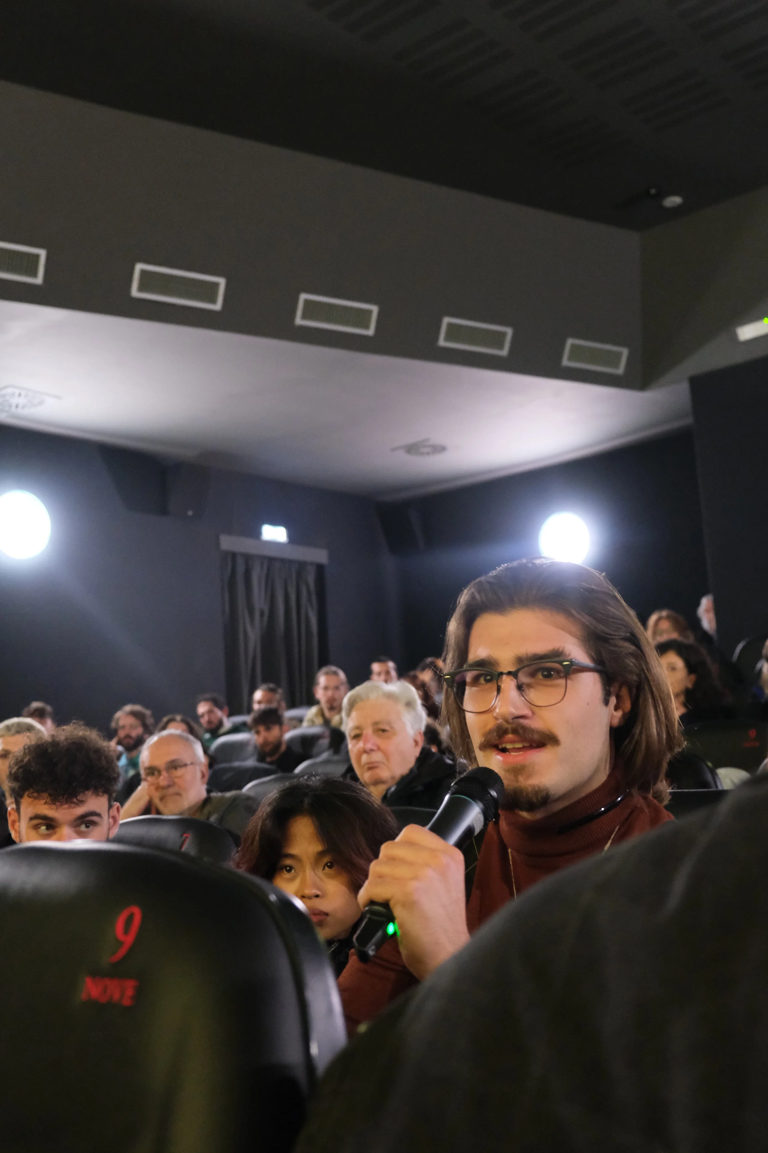 A young man in glasses and a brown turtleneck shirt holding a microphone in a dark auditorium, surrounded by an audience of diverse adults.