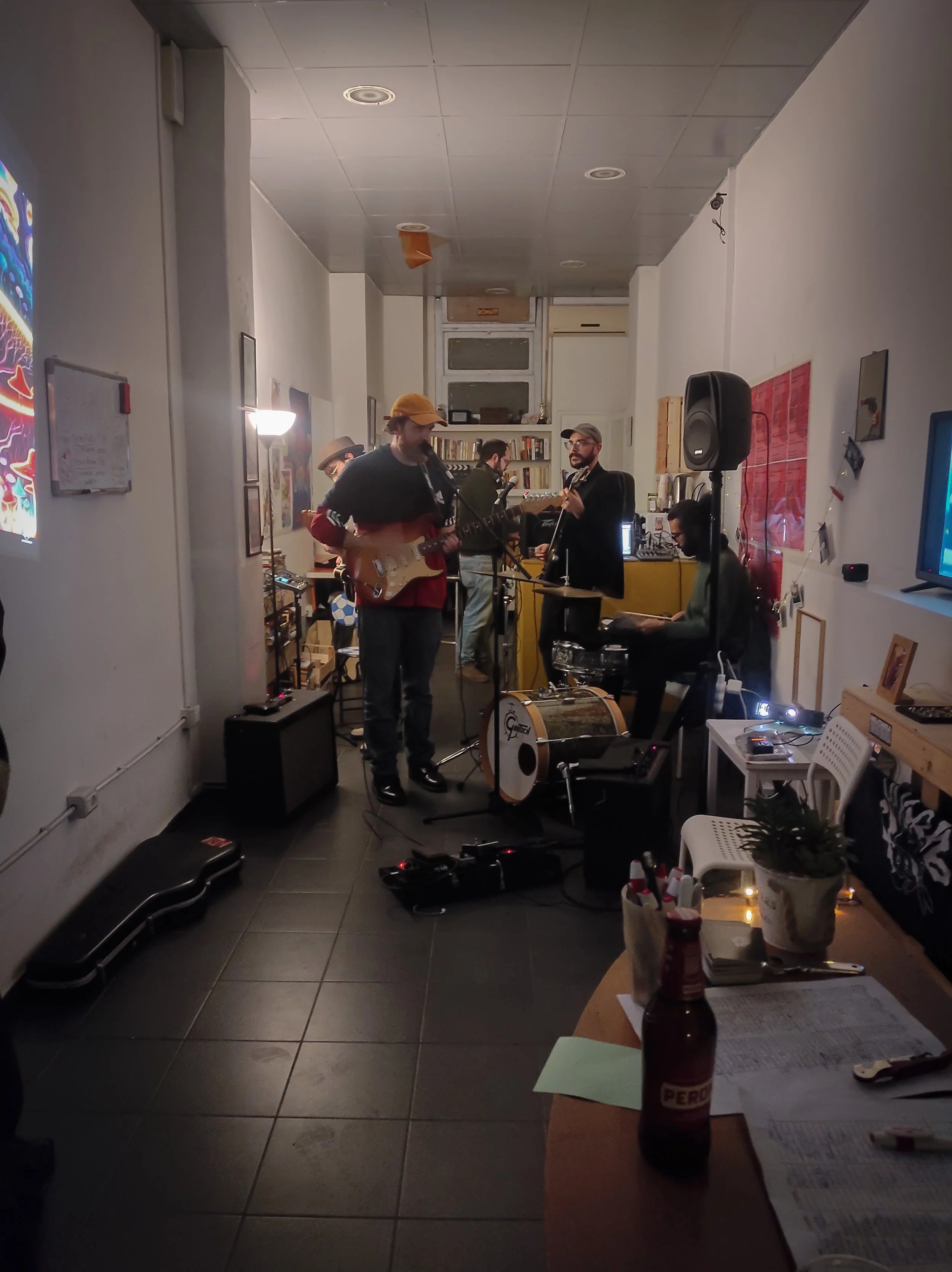 A band performing live music in an indoor setting with four musicians playing guitars and keyboard, surrounded by equipment and audience members.