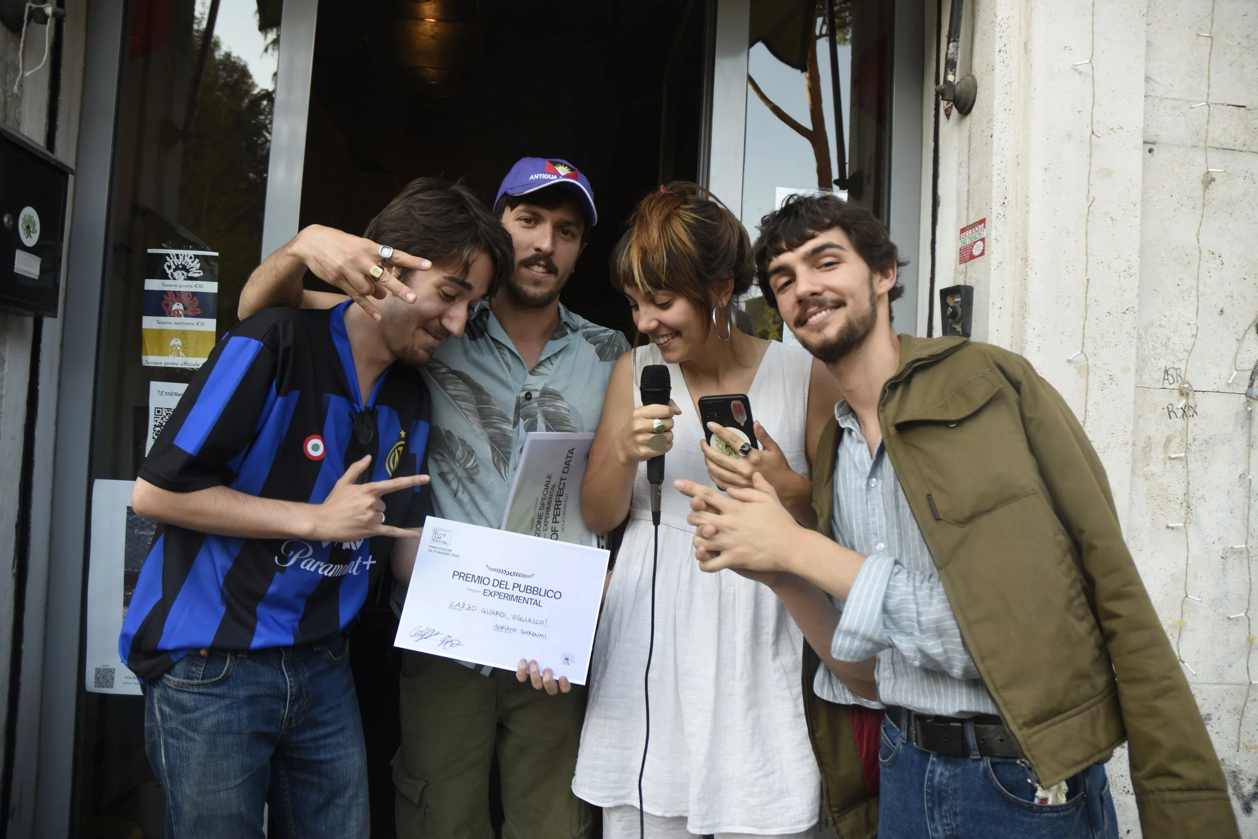 Group of five young adults standing outside a building, celebrating with a certificate and taking selfies. One person holds a microphone, another has a phone, and they are smiling and making gestures.