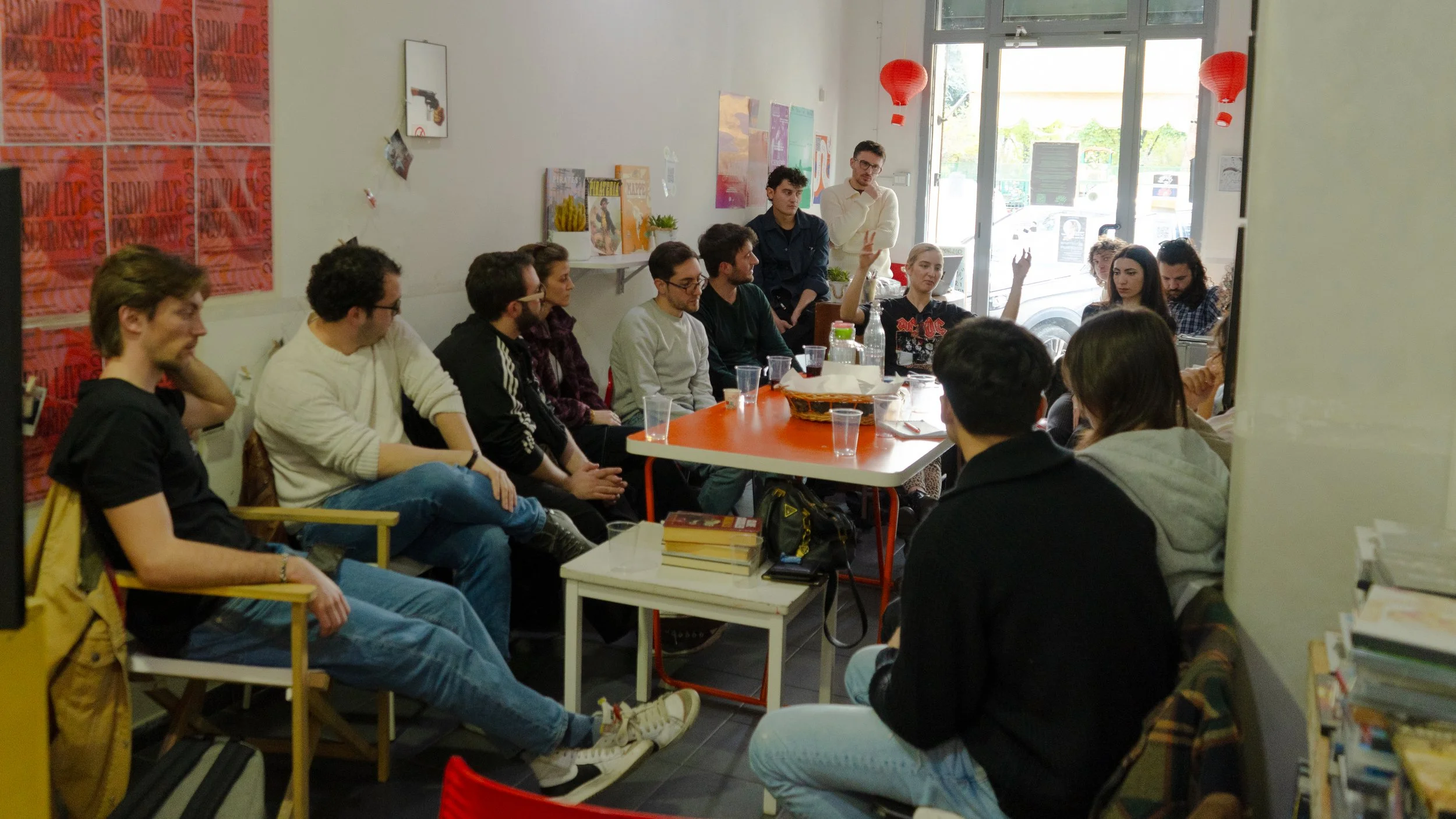 A group of people sitting around a table in a cozy cafe, participating in a meeting or discussion, with some raising hands. The cafe is decorated with red paper lanterns, books, and artwork on the walls, and sunlight coming through large glass doors.