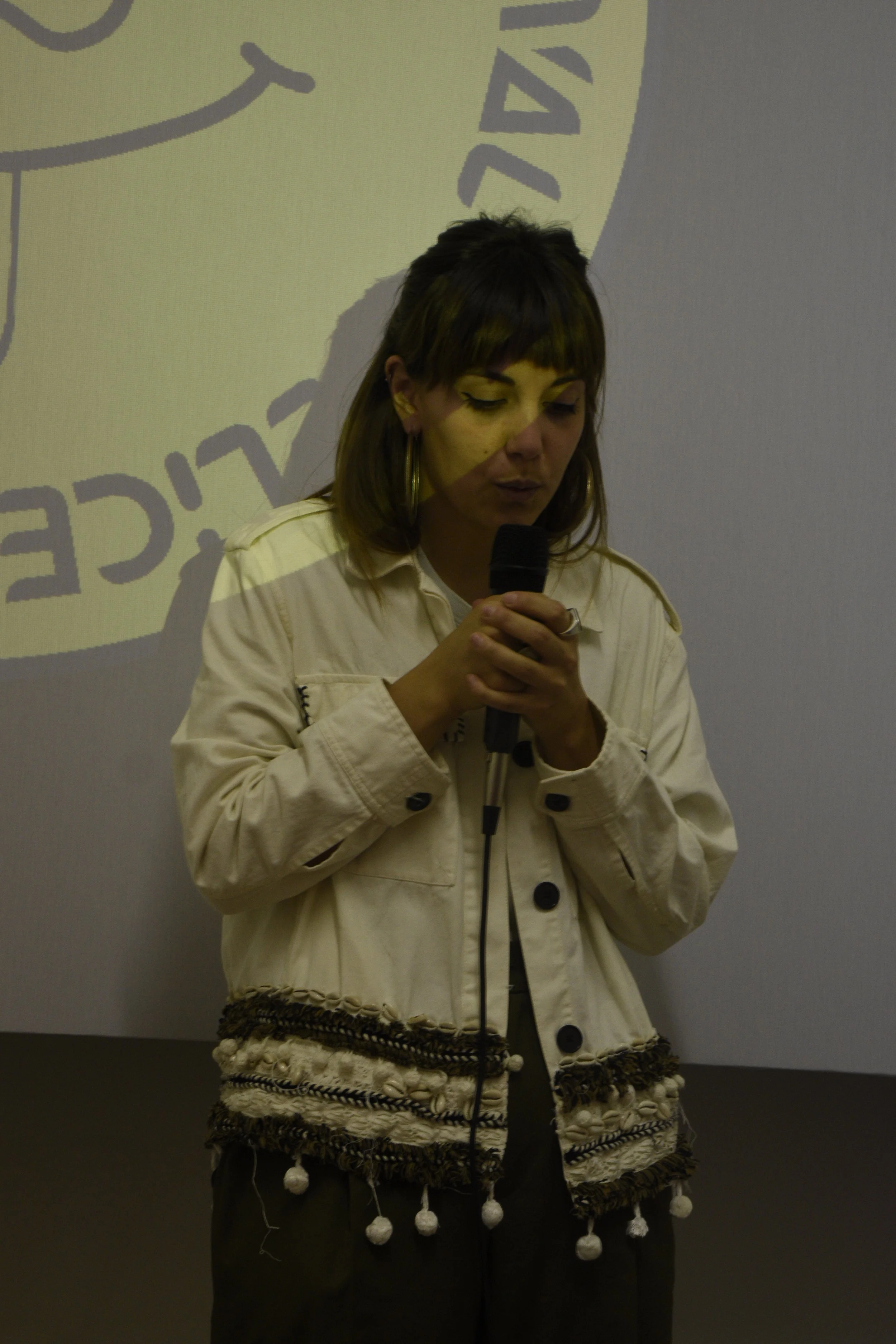 A young woman with brown hair, wearing a cream-colored jacket with black buttons and decorative pom-poms, stands with her eyes closed, holding a microphone close to her mouth, in front of a gray wall with a large logo or graphic partially visible in 