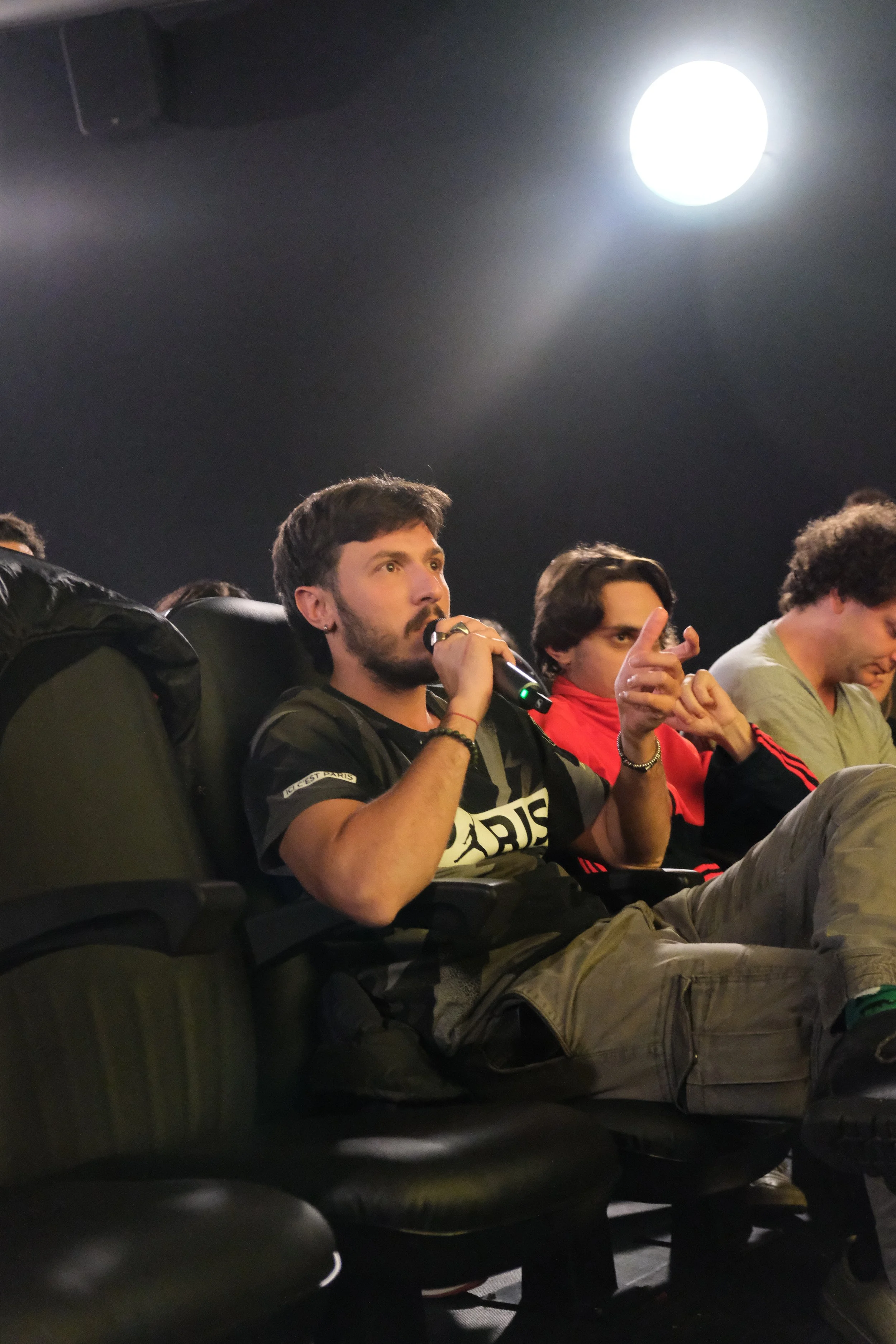 Young man with beard and dark hair sitting in theater, holding microphone and speaking, wearing a black Paris Saint-Germain T-shirt, with others seated beside him, under bright ceiling light.