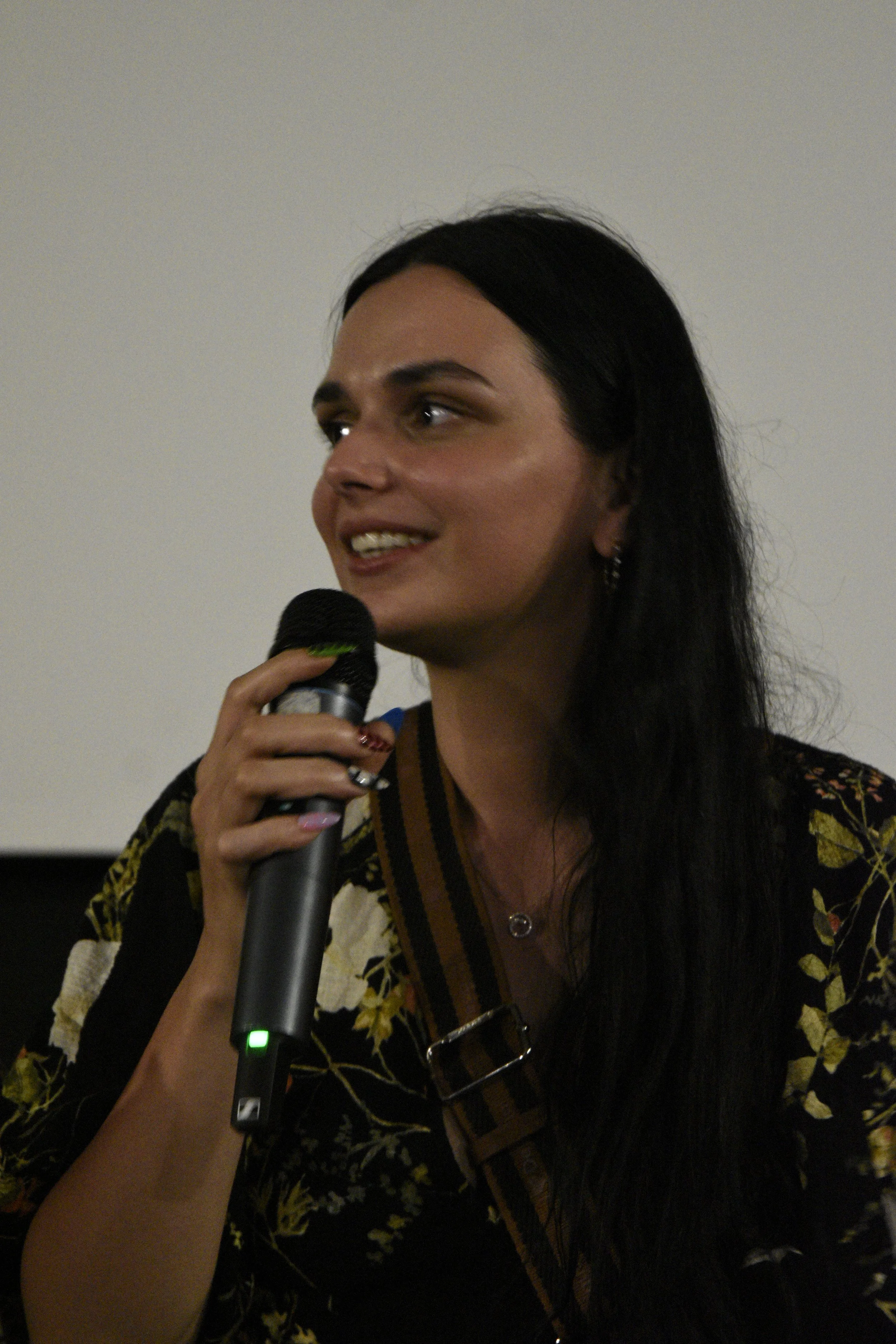 A woman with long black hair speaking into a microphone in an indoor setting.