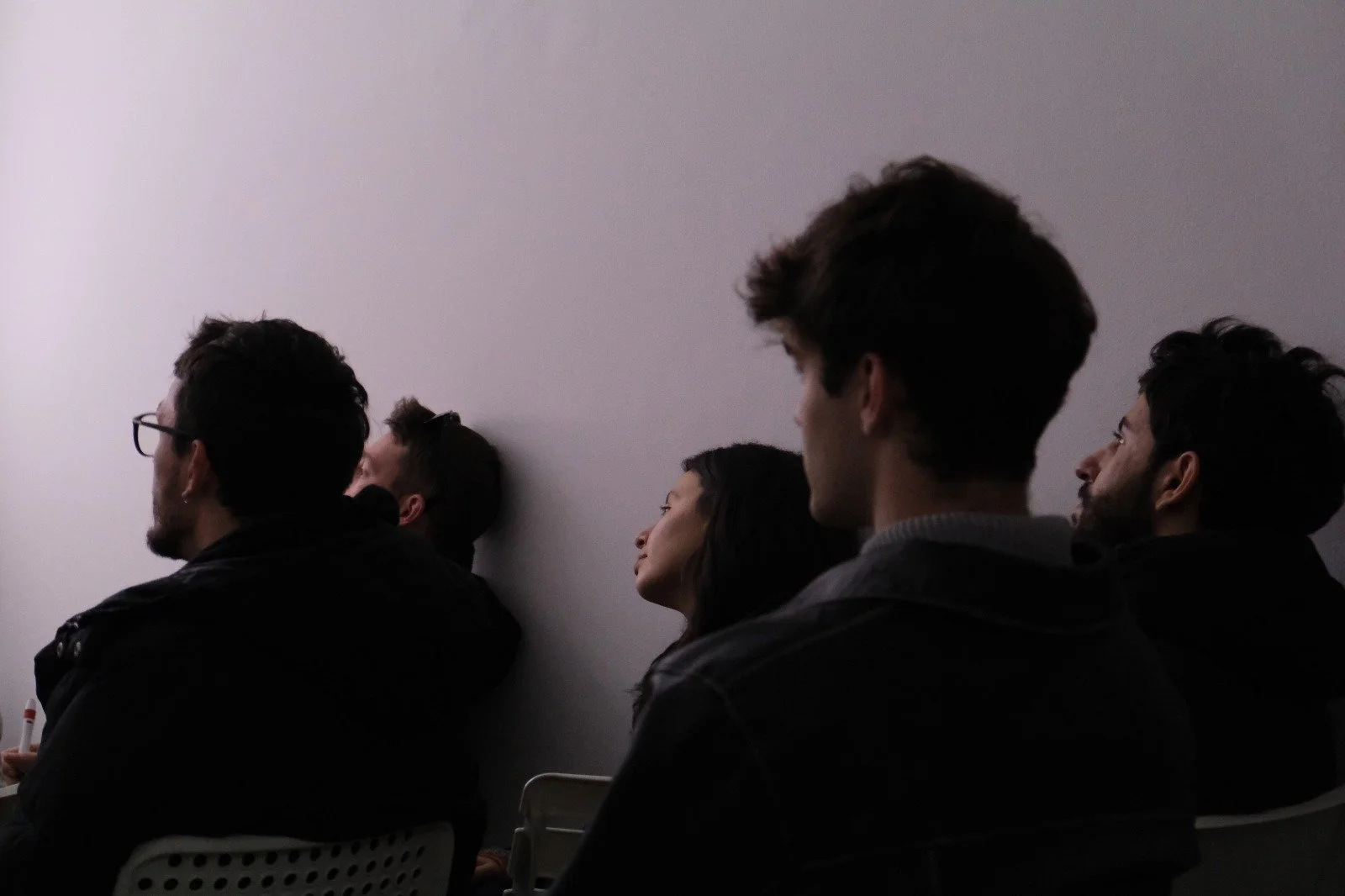 Five people sitting against a plain wall, appearing to listen or observe, in a dimly lit room.