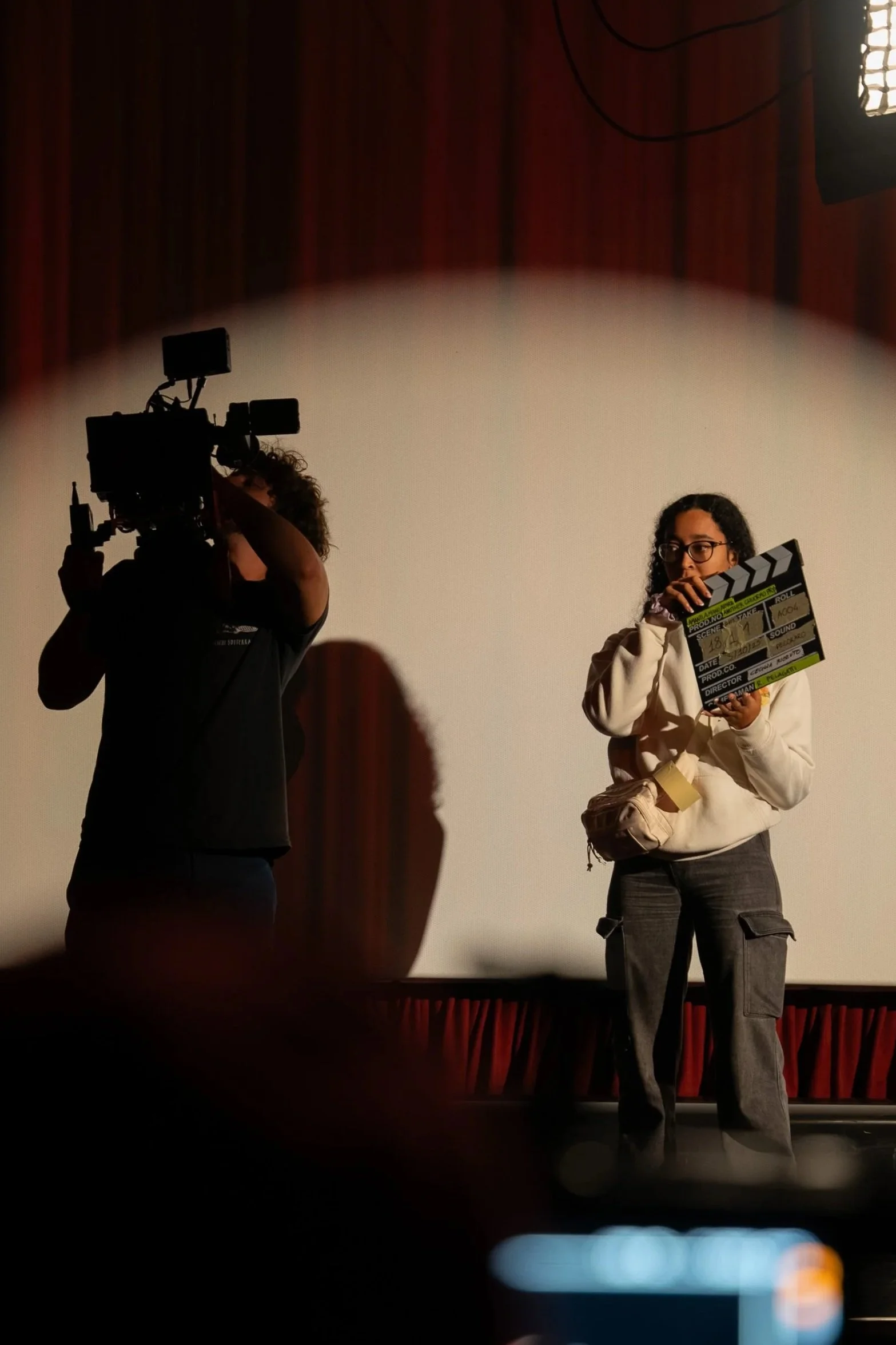 A woman holding a film clapperboard on stage, with a camera operator filming her silhouette against a blank screen.