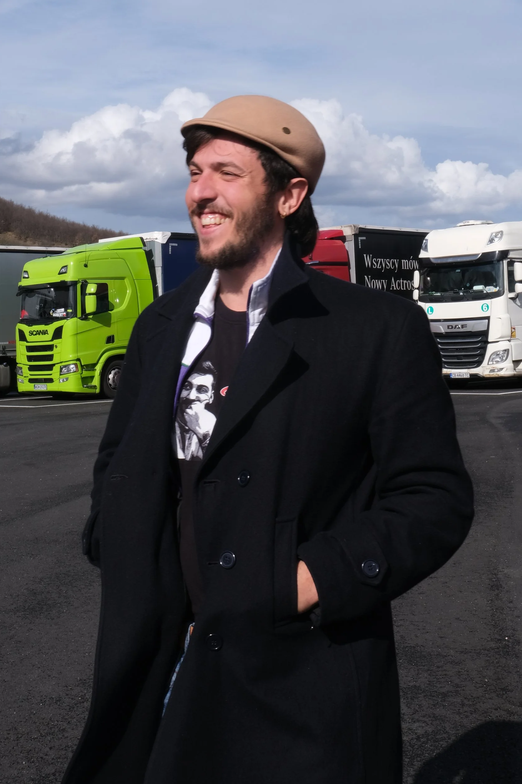 A man with dark hair and a beard smiling and wearing a beige cap and black coat stands outdoors on a cloudy day, with colorful trucks in the background.