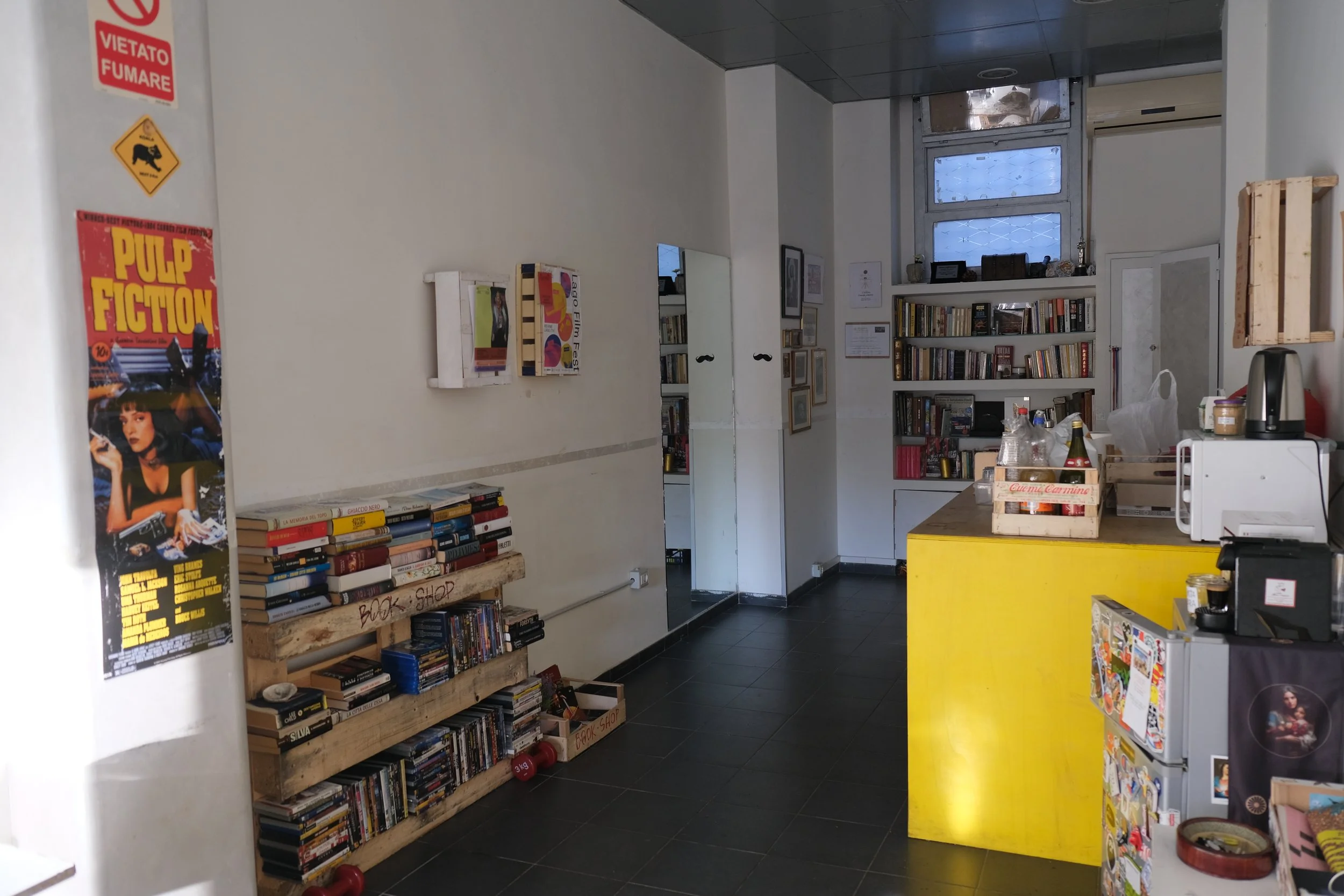 A cozy bookstore or café with bookshelves, a yellow counter, and various decorative items. There's a poster on the left side, and a window at the back letting in natural light.