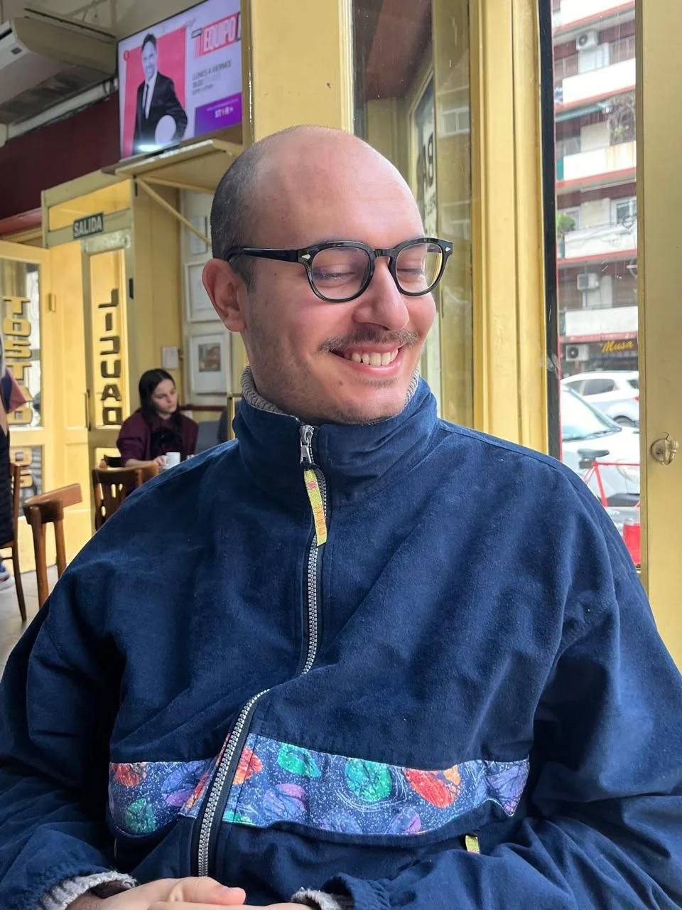 A smiling man with glasses sitting inside a cafe near a large window, wearing a dark blue jacket with a colorful patterned band, with a woman in the background at a table and street view outside.