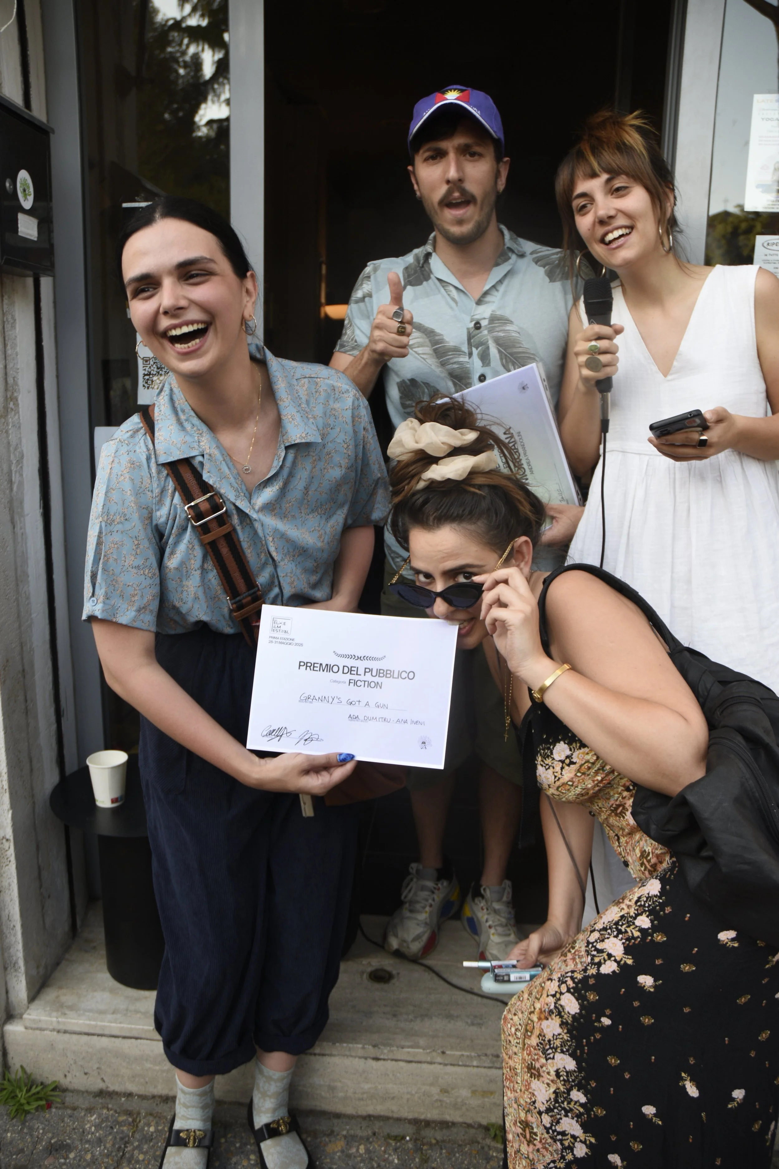 Group of five young people standing outside a building, one woman presenting a certificate for a fiction award, others smiling and engaging with the camera.
