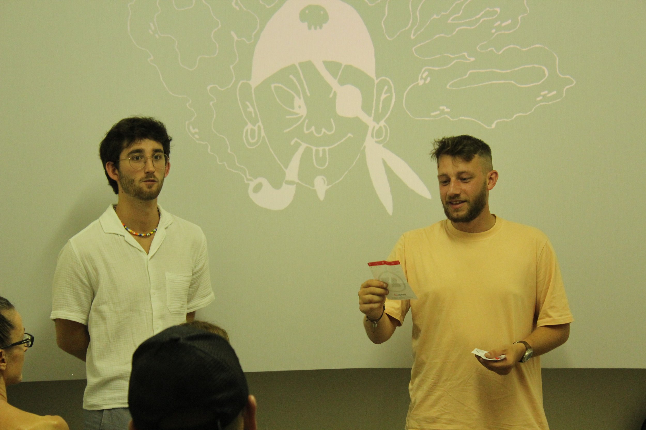 Two men standing in front of a projection of a cartoon drawing of a person with a pipe, earrings, and a howling expression, with a smoke cloud around their head. One man in a light-colored shirt is looking ahead, while the other in a yellow T-shirt i