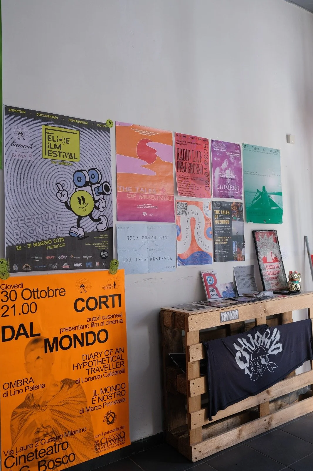 A white wall with various posters advertising films and events, including one for the Greenwich Film Festival and a large orange poster for a film screening. There is a wooden table with pamphlets and a framed picture, a cloth draped over the front of the table, and a Maneki-neko (lucky cat) figurine on the right side of the table.