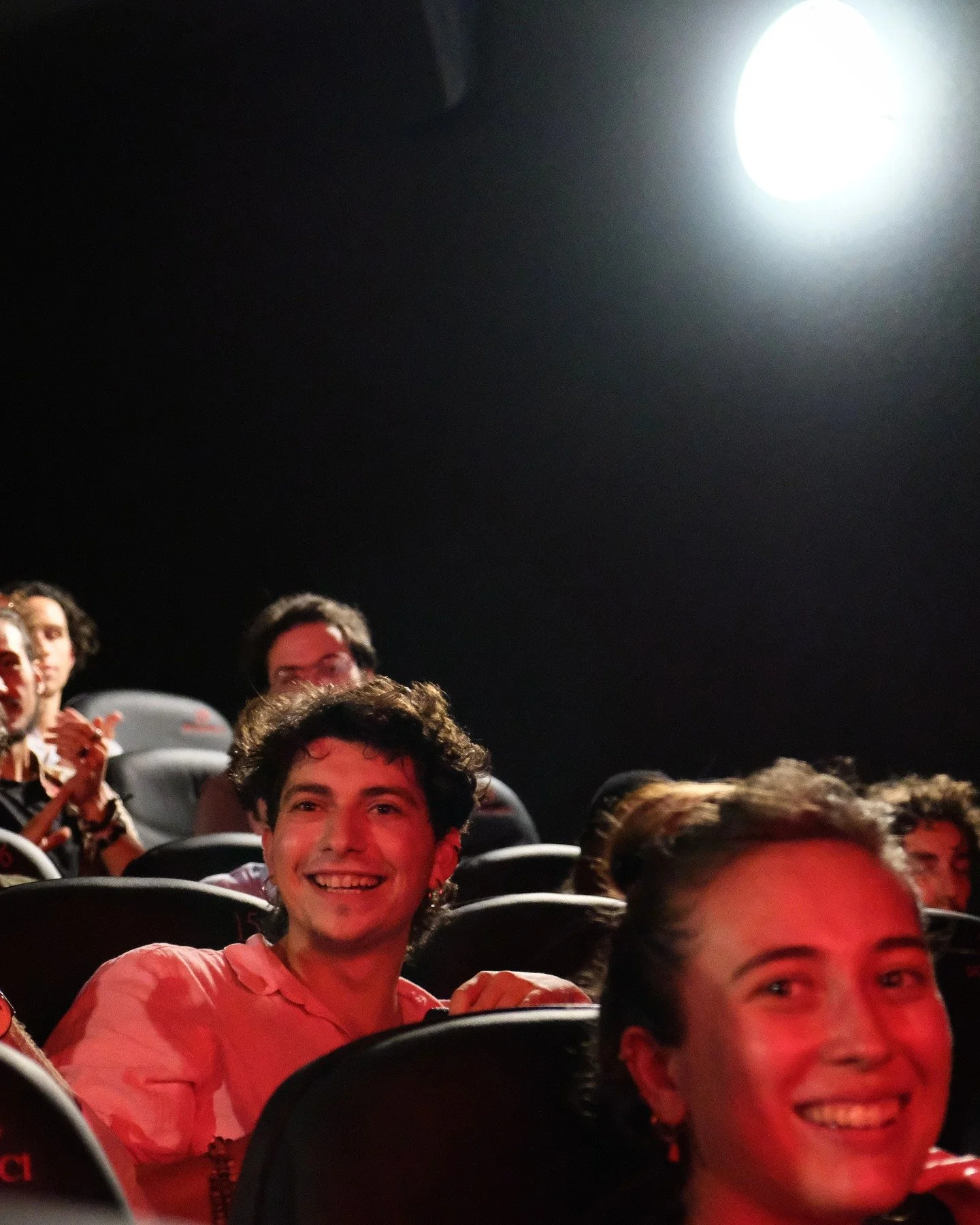 People sitting in a dark theater, smiling and enjoying a movie or event, with some clapping, illuminated by a bright overhead light.
