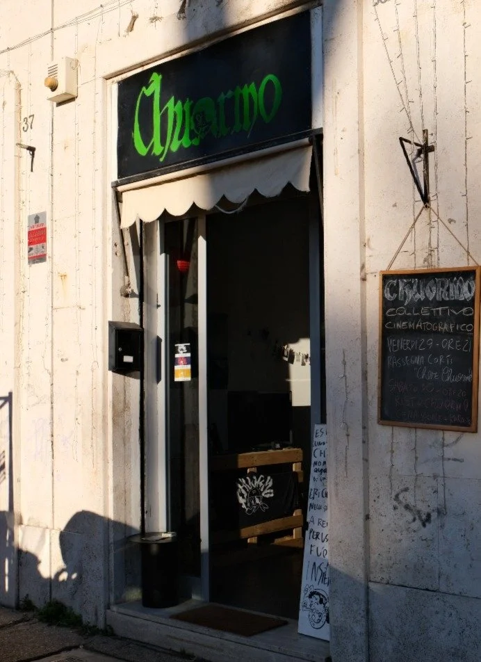 The entrance of a building with a sign that reads "Chavrito," a chalkboard sign with handwritten text in Italian, and a window with a white canopy, suggesting it may be a café or small restaurant.