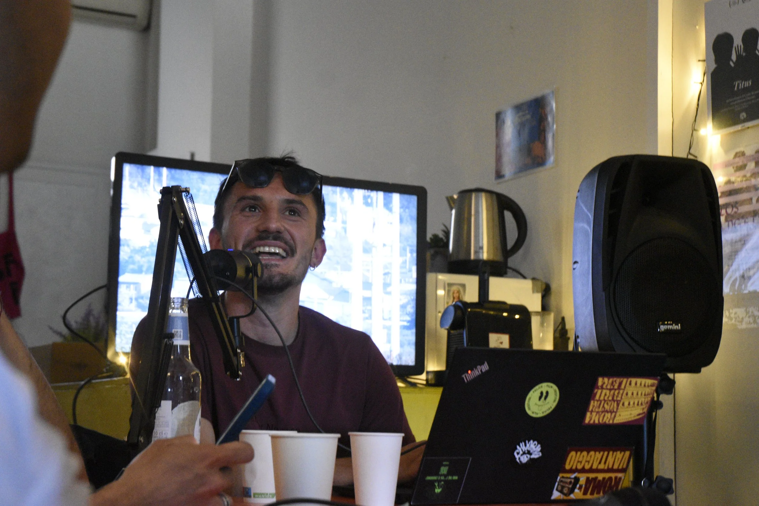 A man with sunglasses on his head and a beard, smiling at a recording session or podcast, with a microphone, a laptop with stickers, a water bottle, and disposable cups in front of him. There is a computer monitor, a speaker, and kitchen appliances v